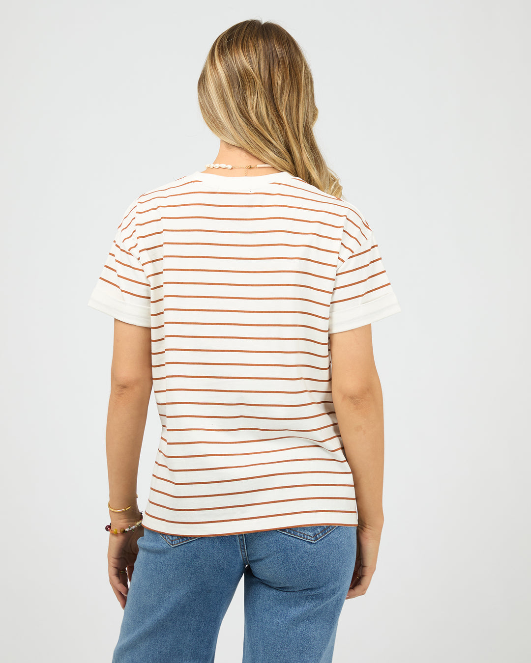 A woman viewed from the back wearing a short-sleeve crewneck t-shirt with thin horizontal rust-orange stripes on an off-white base, paired with blue jeans, against a light gray studio background.