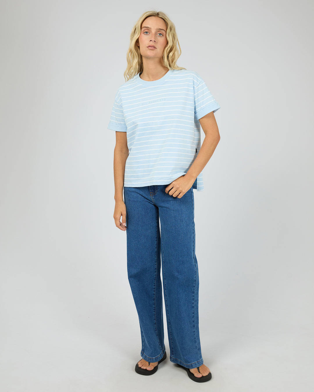 A woman modeling a light blue and white striped short-sleeve t-shirt with wide-leg blue jeans and black sandals, standing against a neutral light gray studio background.