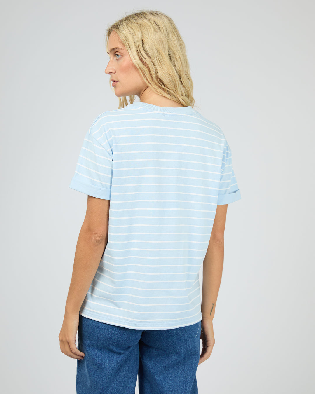 A woman with blonde hair is shown from the back wearing a light blue and white striped short-sleeve t-shirt and blue jeans against a light gray studio background.