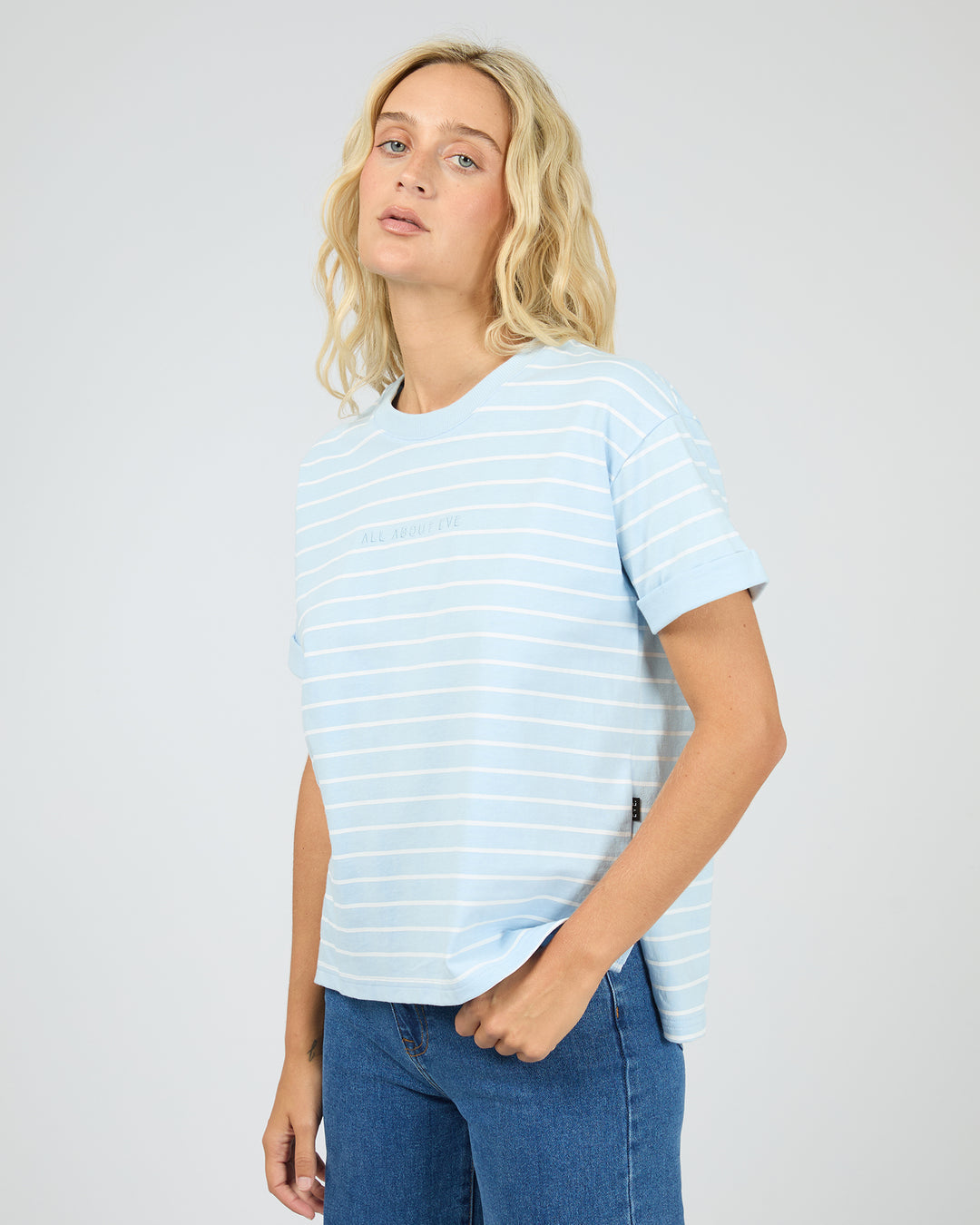 A blonde woman wearing a light blue and white striped short-sleeve t-shirt and blue jeans, standing against a light gray studio background.