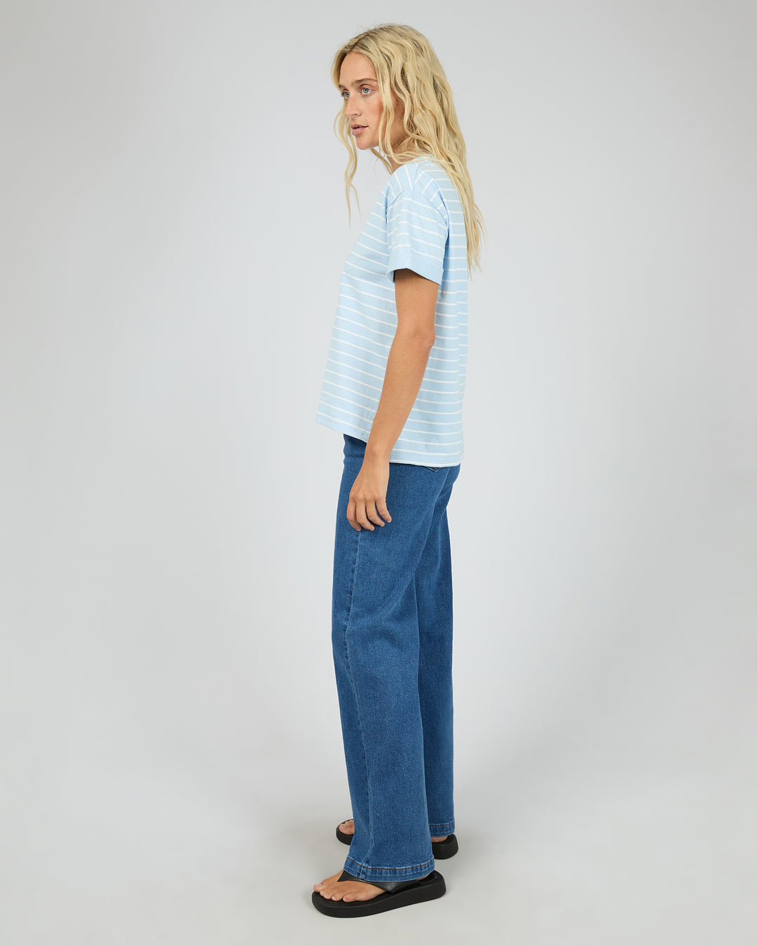 A woman modeling a light blue and white striped short-sleeve t-shirt with wide-leg blue jeans and black sandals, shown from a side profile against a light gray studio background.
