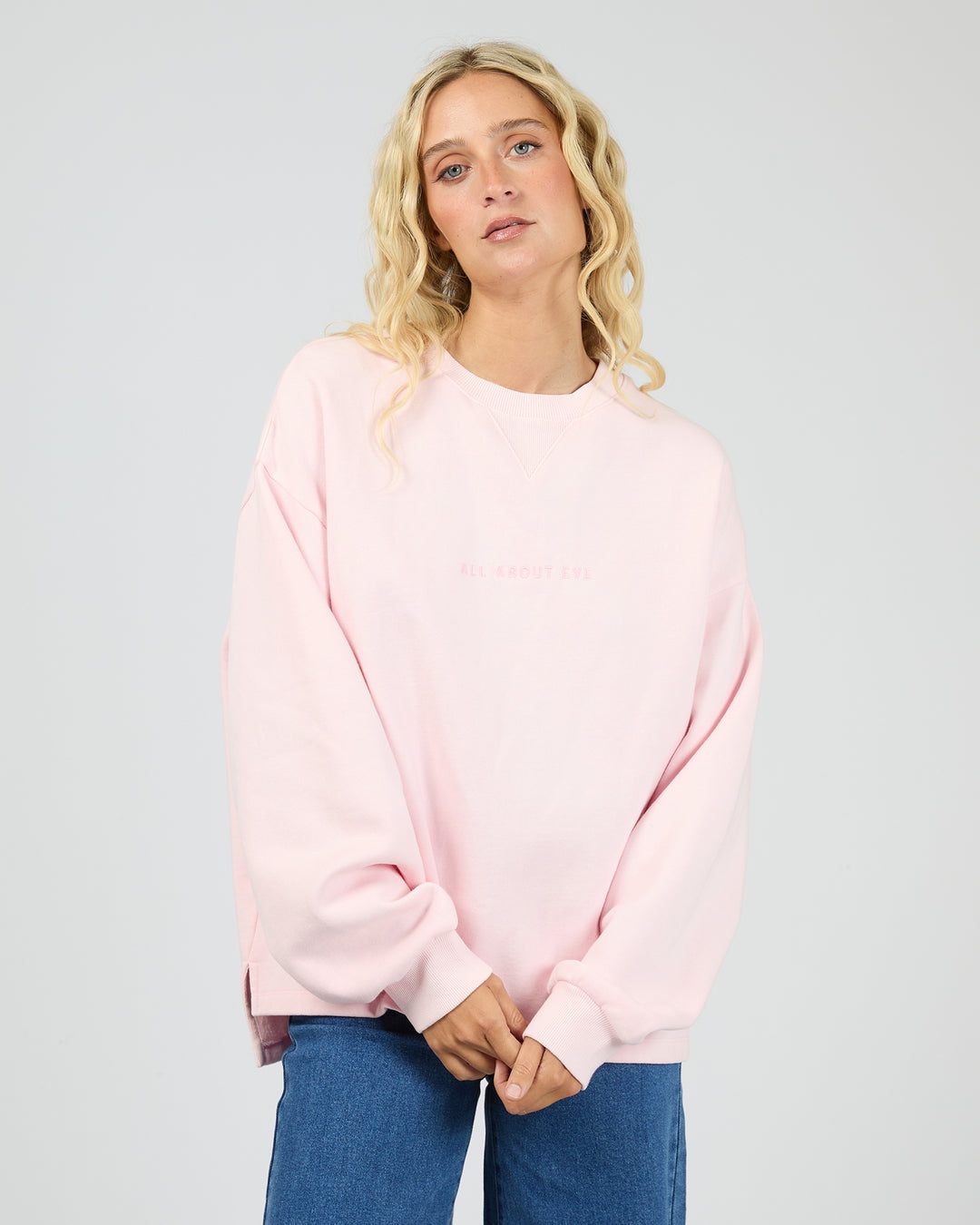 A blonde woman wearing an oversized light pink crewneck sweatshirt with subtle tonal text on the chest, paired with blue jeans, against a plain light gray studio background.