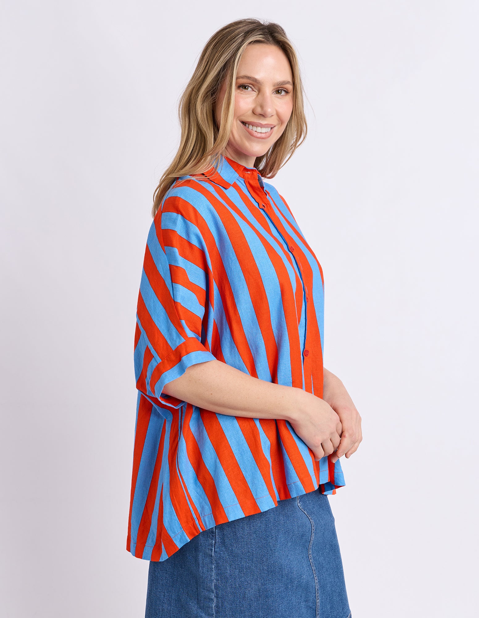 A woman wearing a vibrant blue and orange vertically striped button-up blouse with three-quarter sleeves, paired with a blue denim skirt, photographed against a light gray studio background.