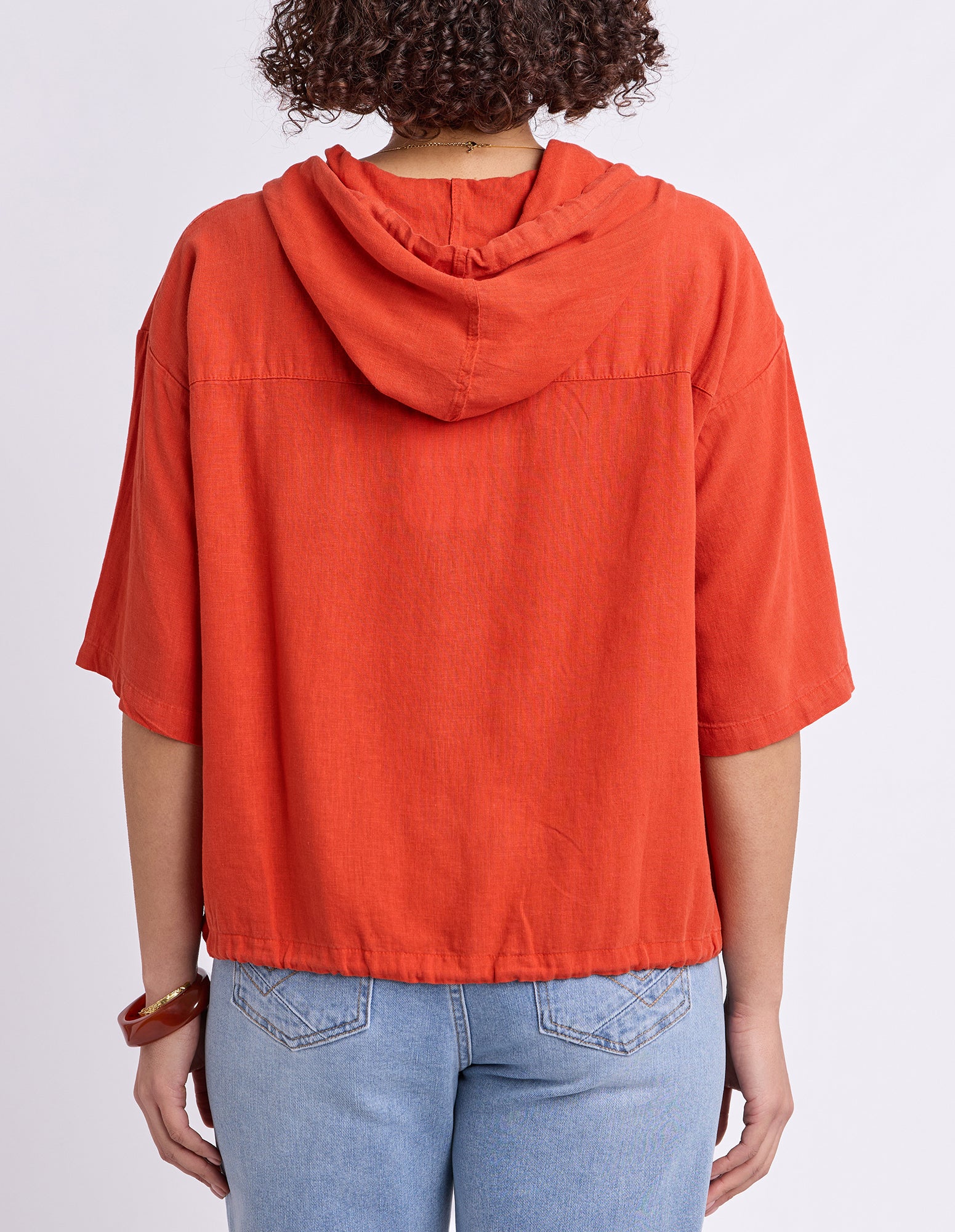 A person shown from the back wearing a bright orange hooded short-sleeve top and light blue jeans against a plain light gray studio background.