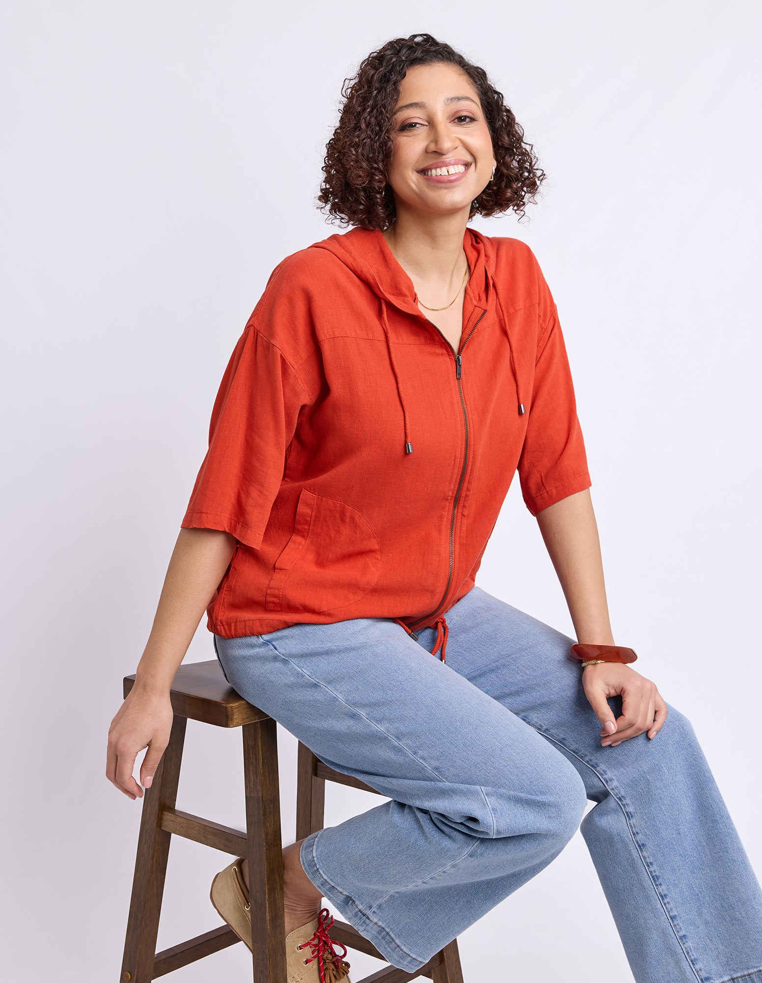 A woman with curly brown hair sits on a wooden stool wearing a rust-orange zip-up hooded jacket and light-wash blue jeans against a plain white studio background.