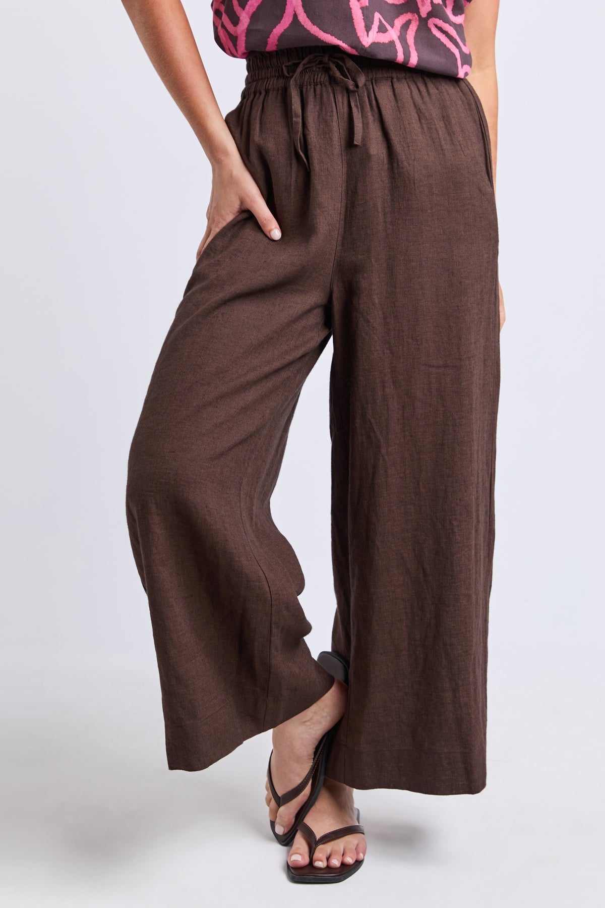 A person wearing loose, wide-leg brown pants with a drawstring waist and side pockets, paired with black strappy sandals against a light gray studio background.