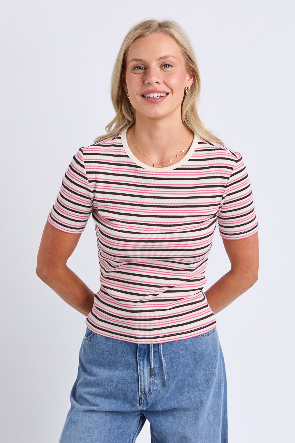A woman modeling a short-sleeve ribbed knit top with horizontal pink, black, and white stripes, paired with blue jeans, against a light gray studio background.