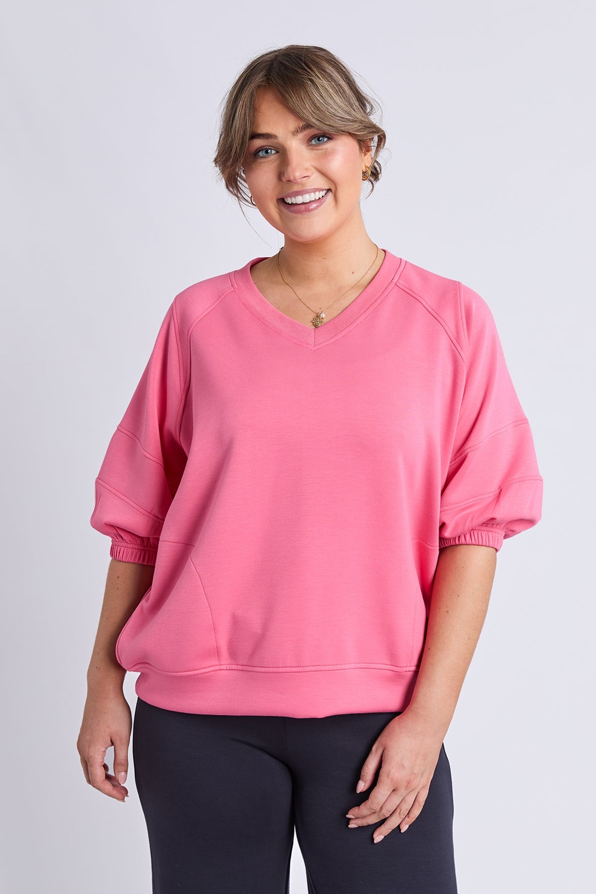 A woman wearing a bright pink V-neck sweatshirt with elbow-length balloon sleeves and elastic cuffs, paired with dark leggings, against a light gray studio background.