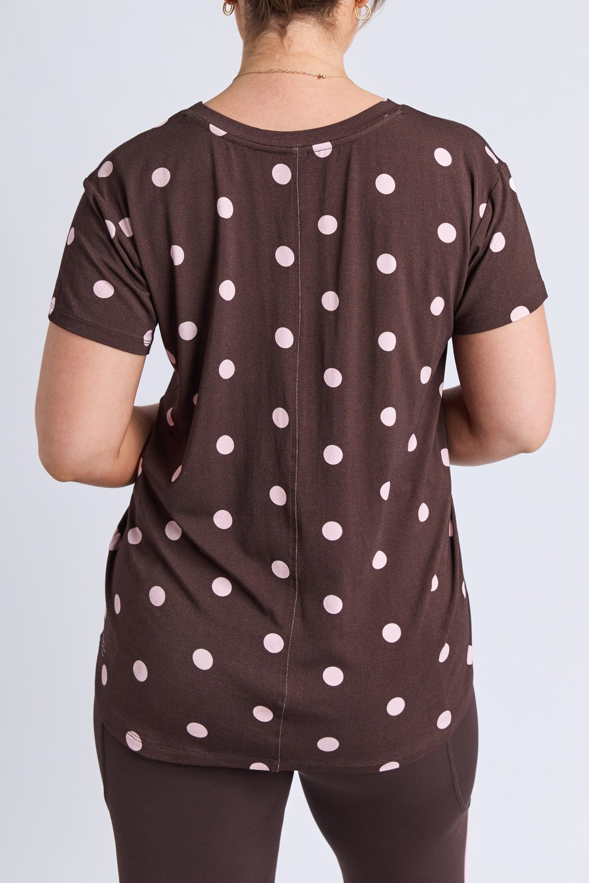 A woman viewed from the back wearing a dark brown short-sleeve top with a white polka dot pattern and dark brown leggings, set against a plain light gray studio background.