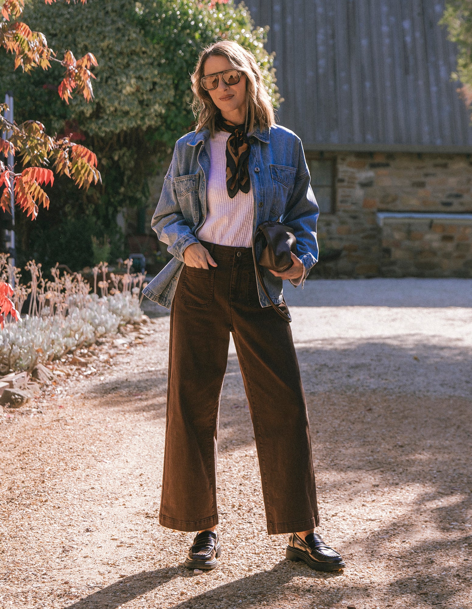 Woman standing outdoors wearing a denim jacket, white shirt, brown pants, and black shoes.
