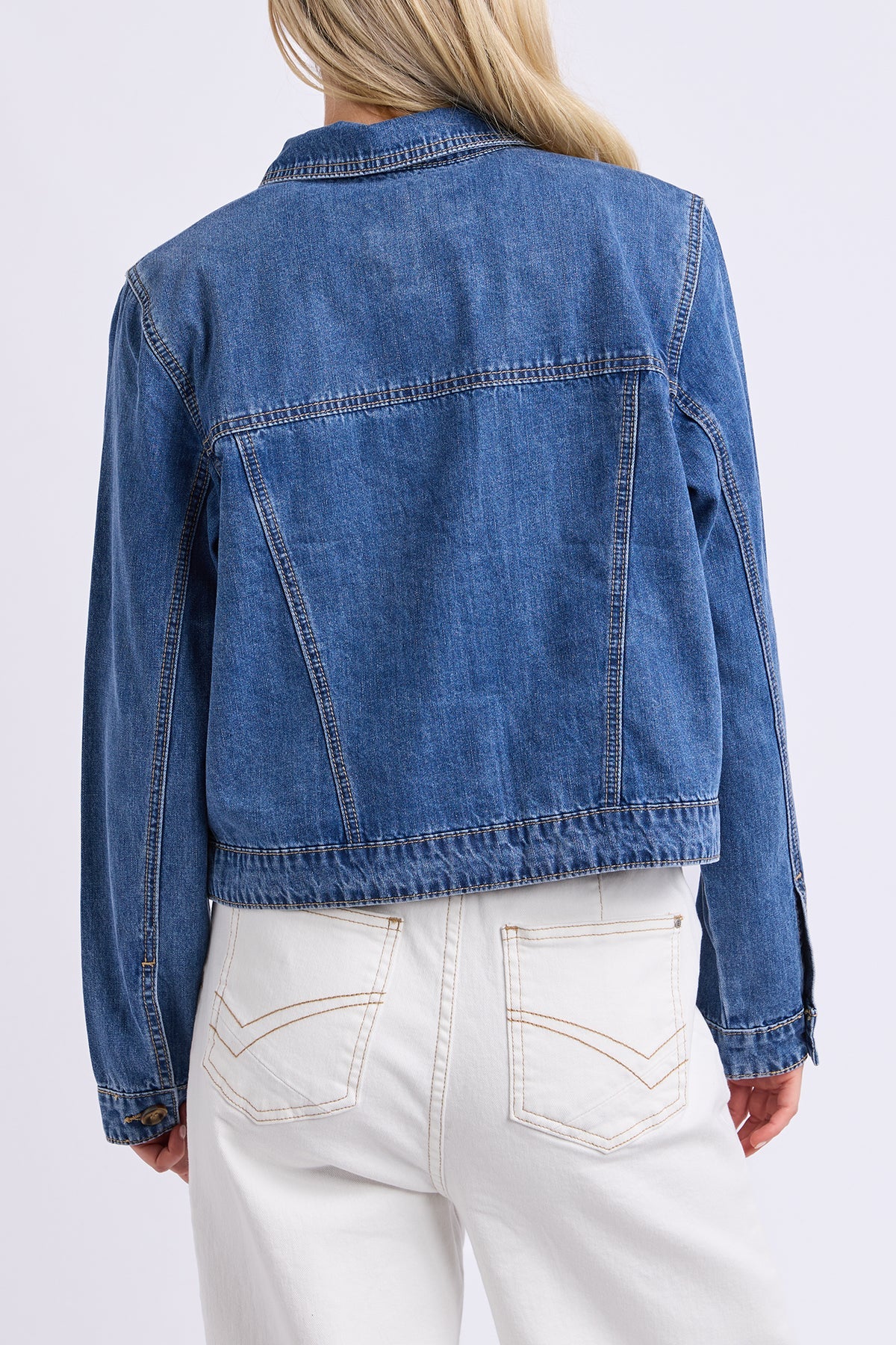 A person shown from the back wearing a cropped blue denim jacket with white pants, photographed against a clean light gray studio background.