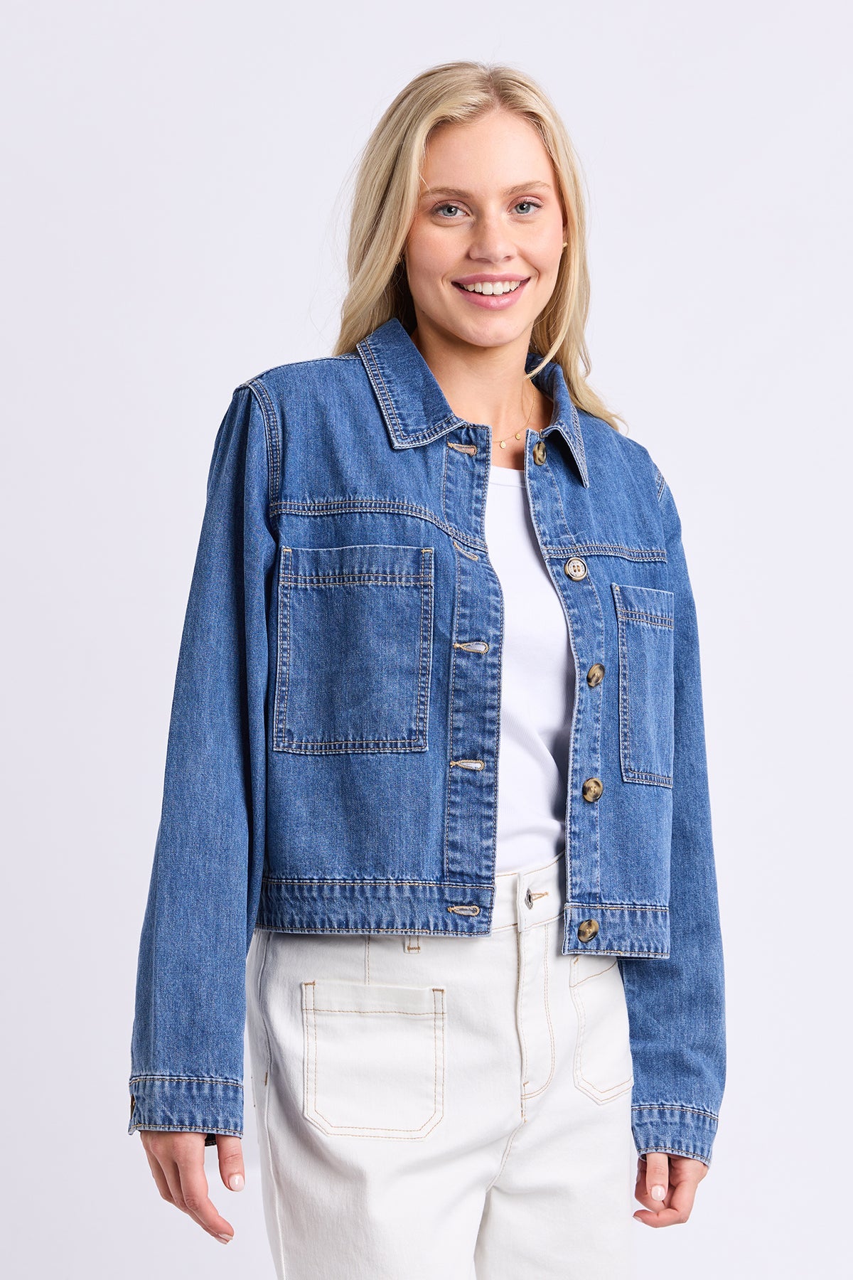 A woman wearing a cropped blue denim jacket over a white top and white pants, standing against a light gray studio background.