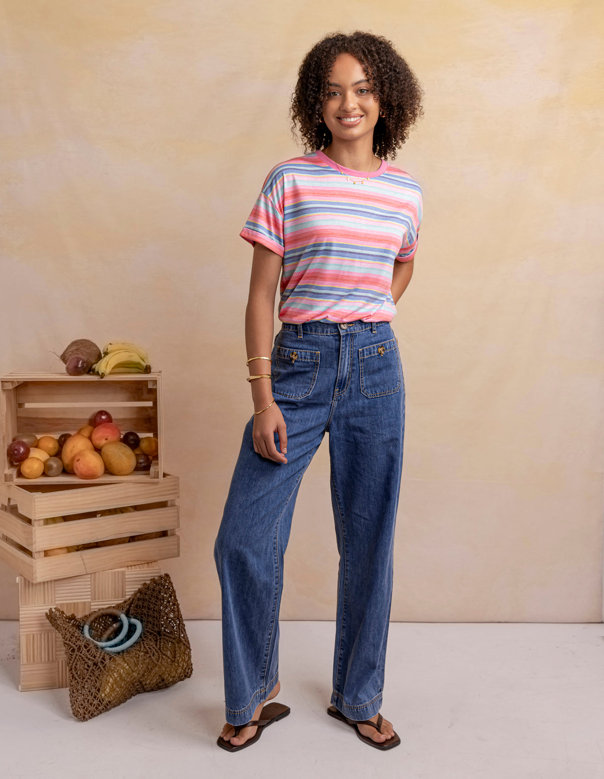 A woman stands in a studio wearing a short-sleeve striped t-shirt in pink, blue, and white, paired with high-waisted wide-leg blue jeans and brown sandals. To her left are wooden crates holding fruit and a woven bag, set against a warm beige textured backdrop.