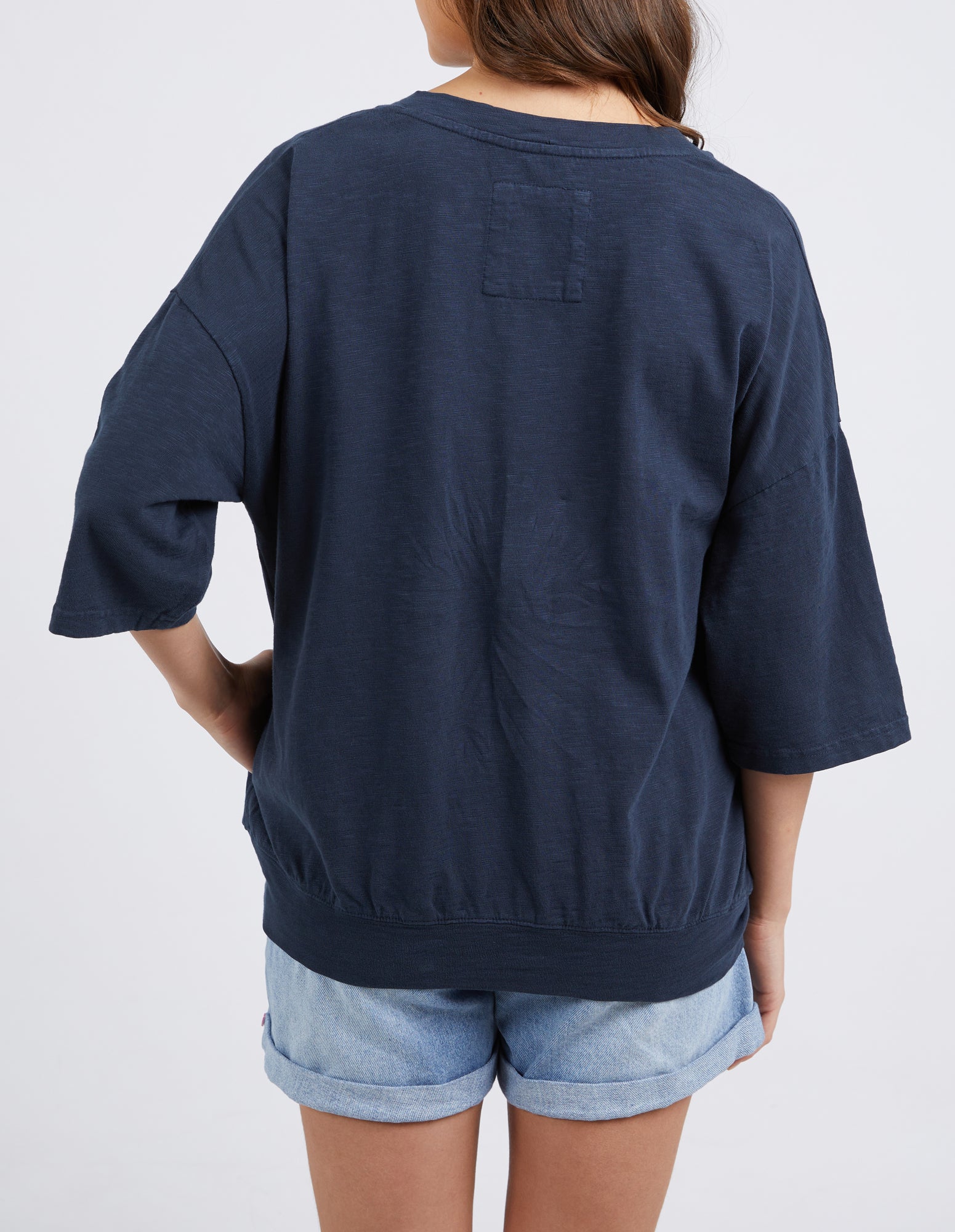 A person shown from the back wearing an oversized dark navy blue short-sleeve top and light blue denim shorts against a plain light gray studio background.