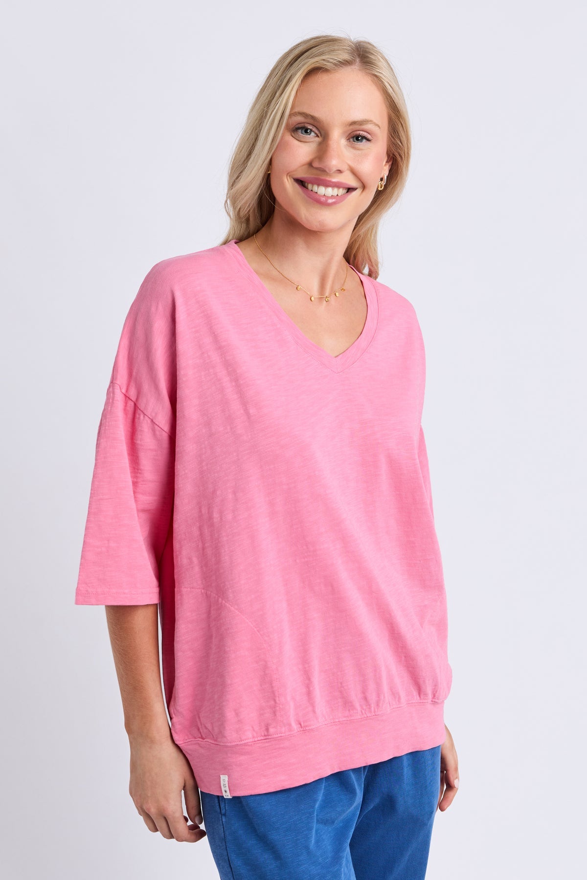 A smiling blonde woman wearing a pink V-neck knit top with three-quarter sleeves and blue pants, standing against a light gray studio background.