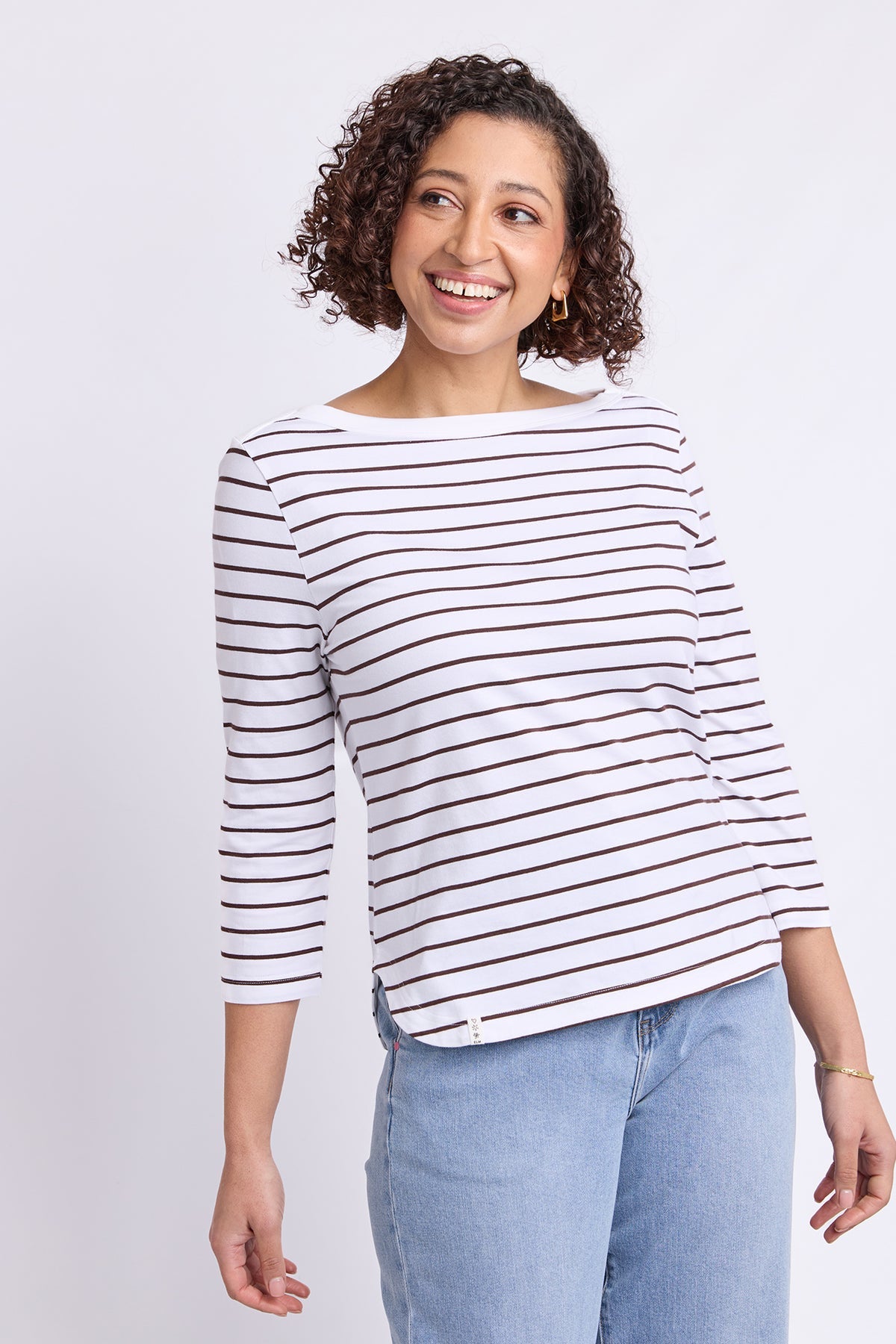 A smiling woman with curly brown hair wearing a white three-quarter sleeve top with thin dark brown horizontal stripes and light blue jeans, standing against a light gray studio background.