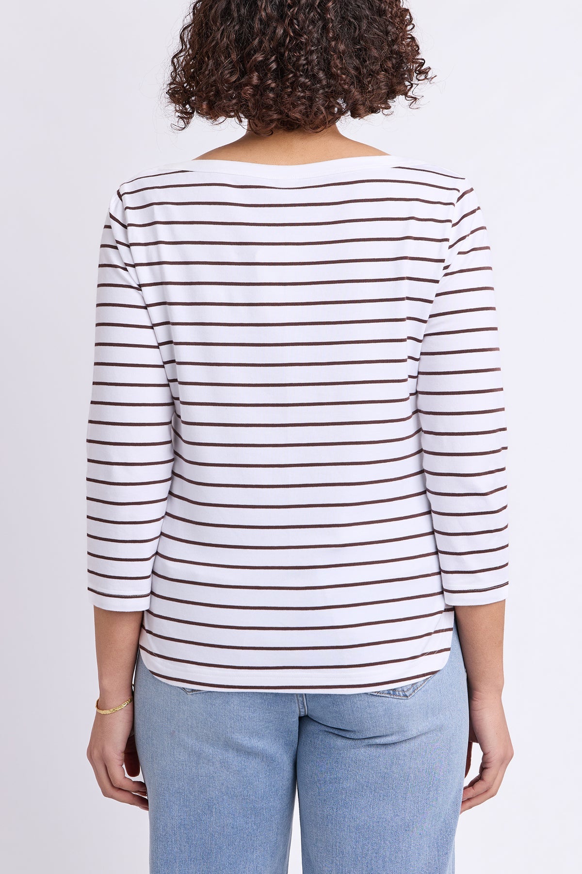 A woman viewed from the back wearing a white long-sleeve top with thin brown horizontal stripes and light blue jeans, against a plain light gray studio background.