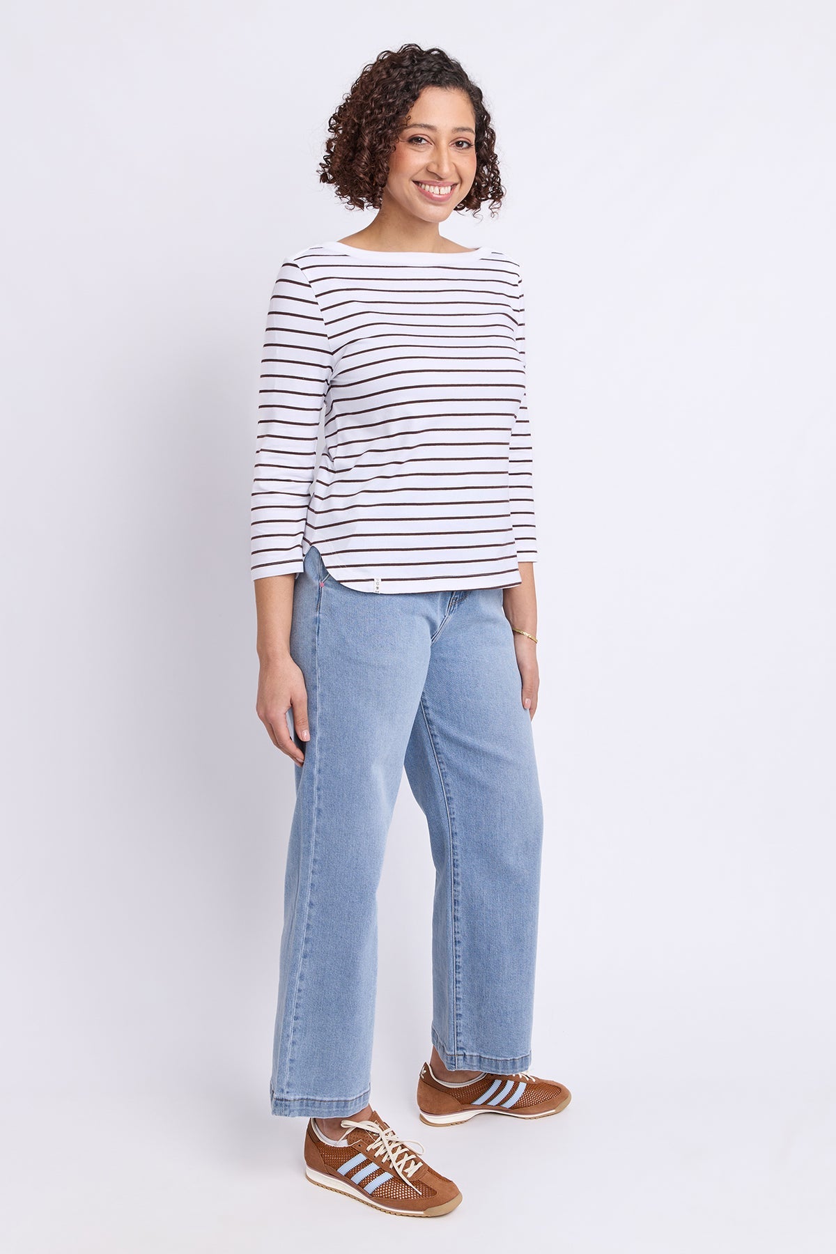 A woman modeling a white long-sleeve top with thin dark horizontal stripes, paired with light blue wide-leg jeans and brown sneakers, against a plain white studio background.