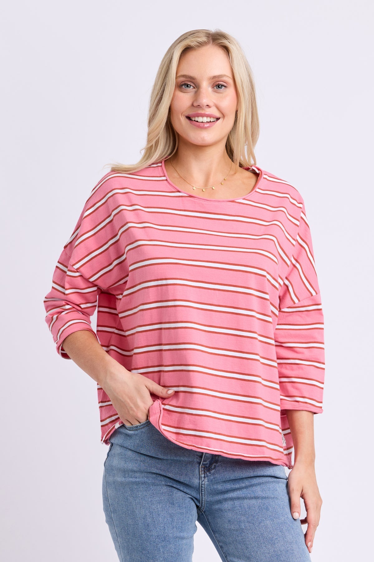 A woman modeling a pink long-sleeve top with thin white and beige horizontal stripes, paired with blue jeans, against a light gray studio background.