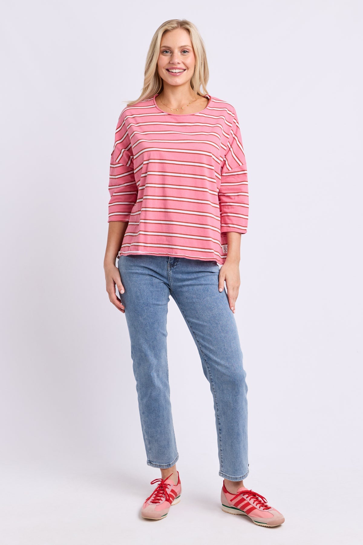 A woman modeling a pink and white striped long-sleeve top, paired with light blue jeans and pink sneakers, against a plain light gray studio background.