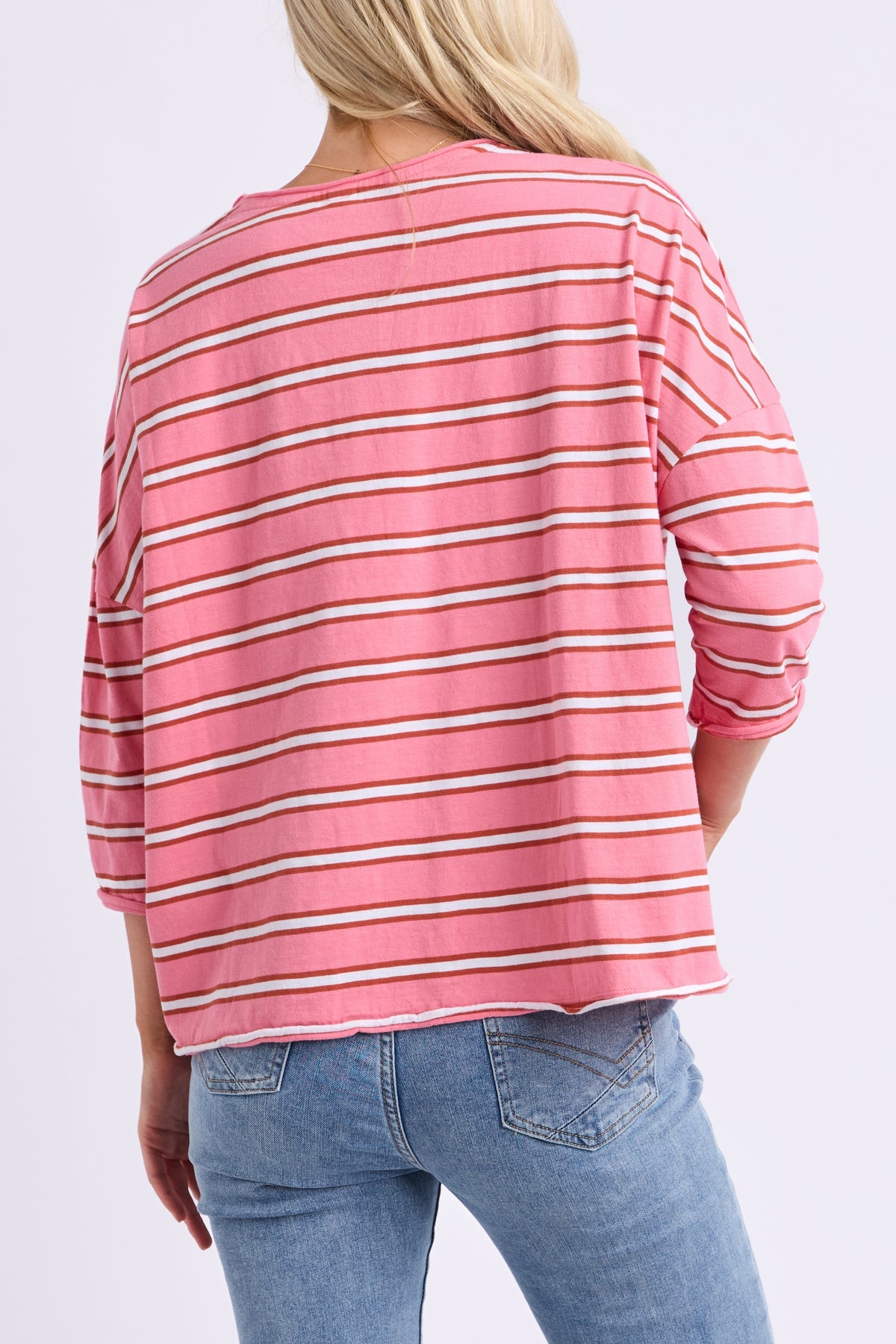 A woman shown from the back wearing a pink long-sleeve top with thin white and beige horizontal stripes, paired with blue jeans, against a light gray studio background.