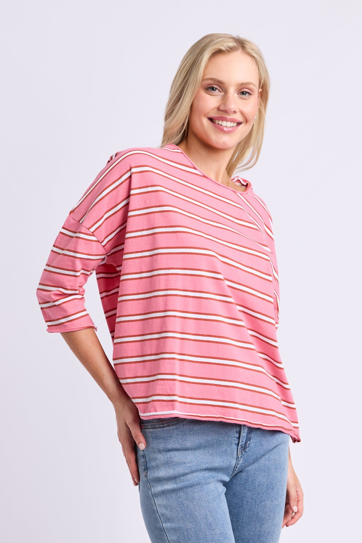A smiling blonde woman wearing a pink three-quarter sleeve top with thin white and orange horizontal stripes, paired with blue jeans, against a light gray studio background.