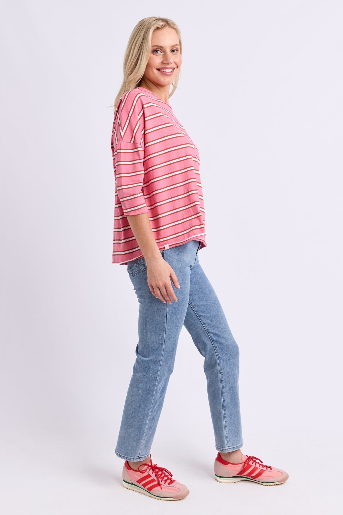 A woman modeling a pink and white striped three-quarter sleeve top, paired with light blue jeans and pink sneakers, against a light gray studio background.