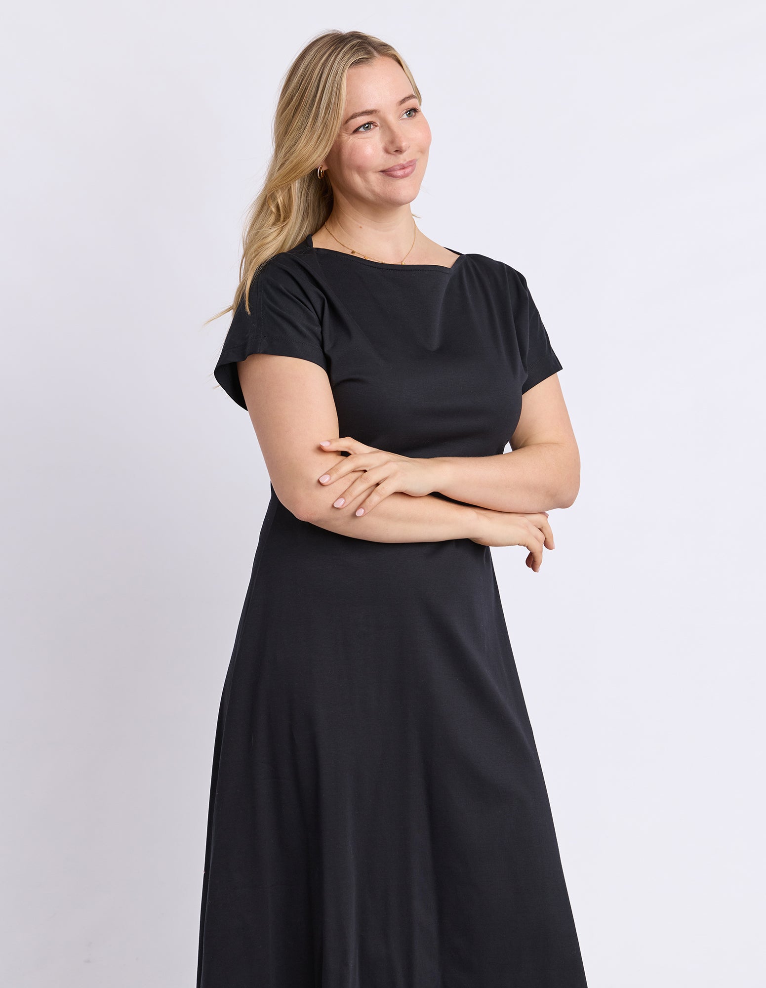 A woman with blonde hair wearing a black short-sleeve maxi dress, standing with her arms crossed against a light gray studio background.