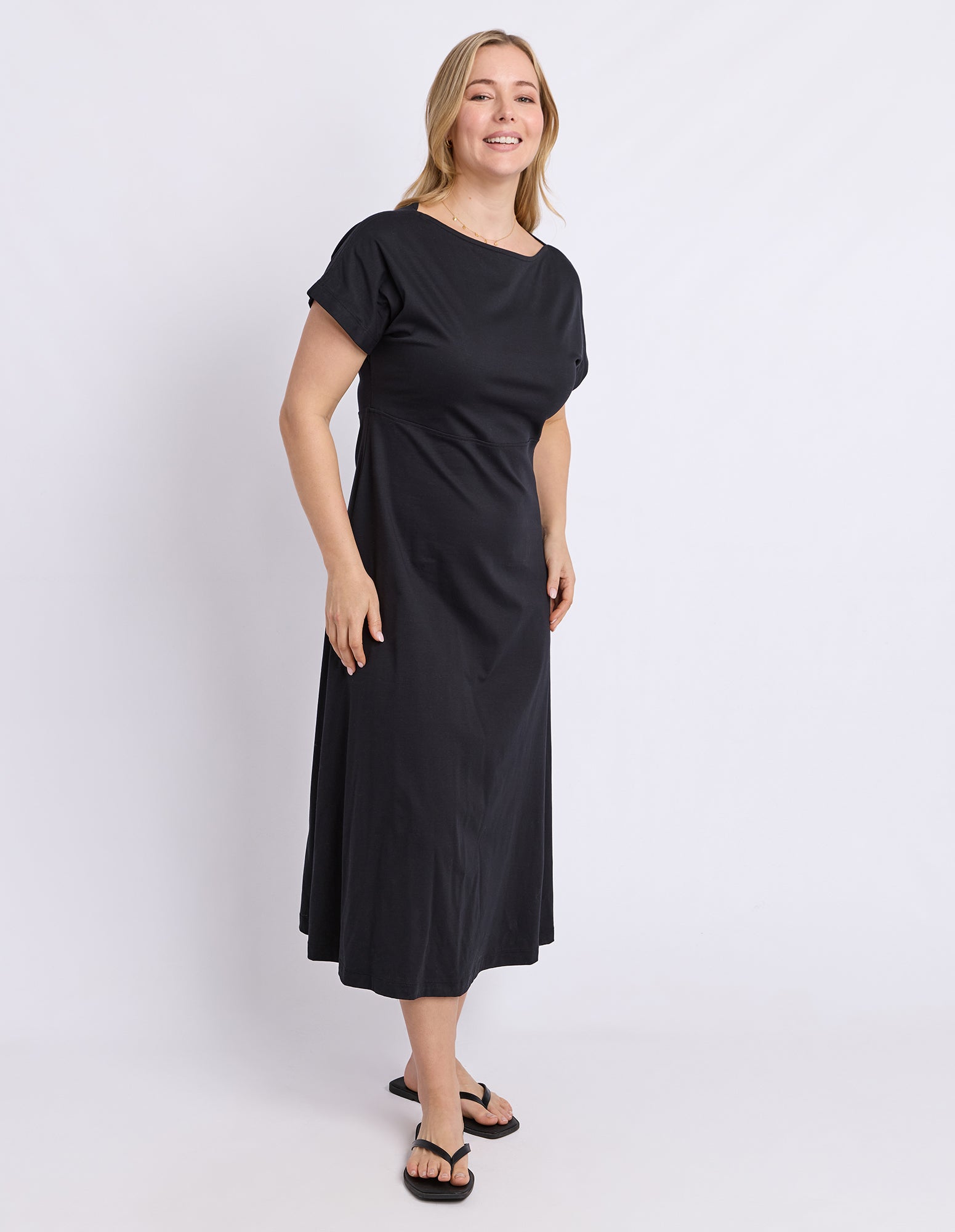 A woman modeling a black short-sleeve midi dress with a boat neckline, photographed against a plain light gray studio background.