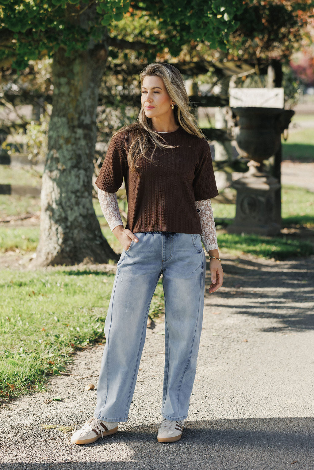 Woman wearing a brown sweater and light blue jeans standing on a path with trees in the background