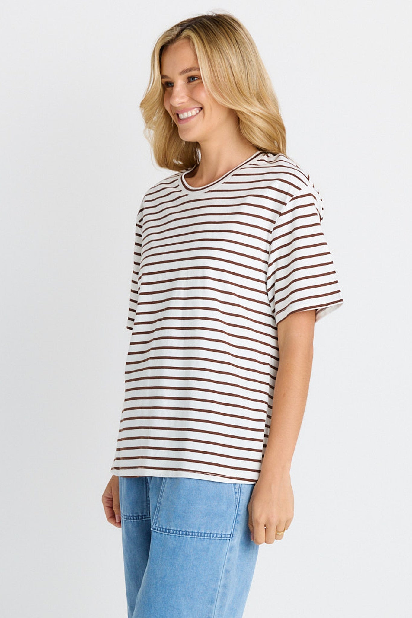 A woman wearing a short-sleeve crewneck t-shirt with thin brown horizontal stripes on an off-white base, paired with light blue jeans, photographed against a light gray studio background.