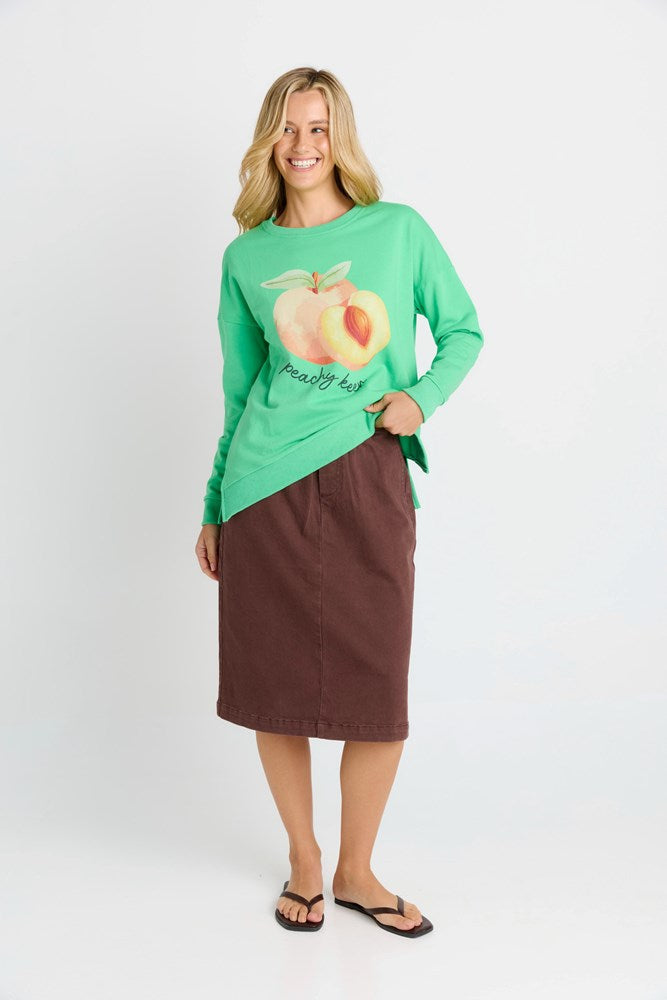 Woman wearing a green long-sleeve shirt with a peach graphic and brown skirt on a white background