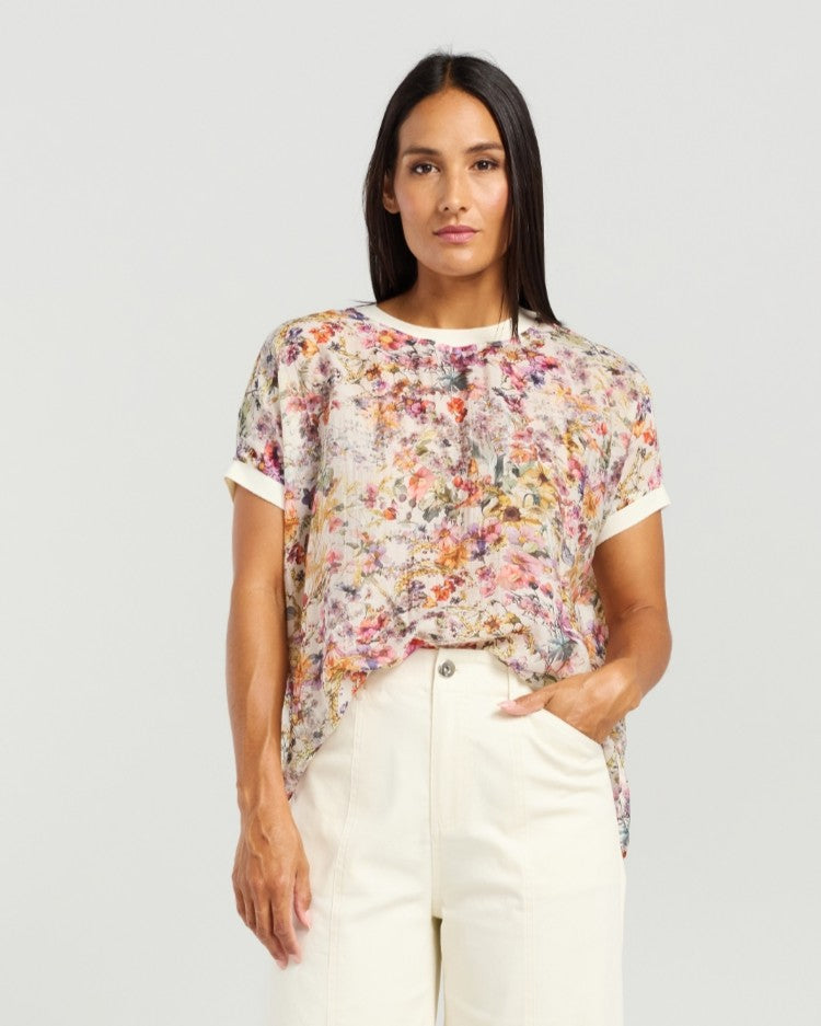 A woman modeling a short-sleeve t-shirt with a dense multicolor floral print over a white base, paired with cream-colored pants, against a solid light gray background.