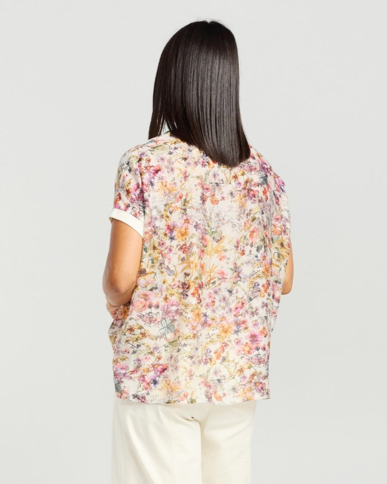 A woman shown from the back wearing a short-sleeve floral blouse with a cream base and multicolored floral print, paired with light beige pants, against a light gray studio background.
