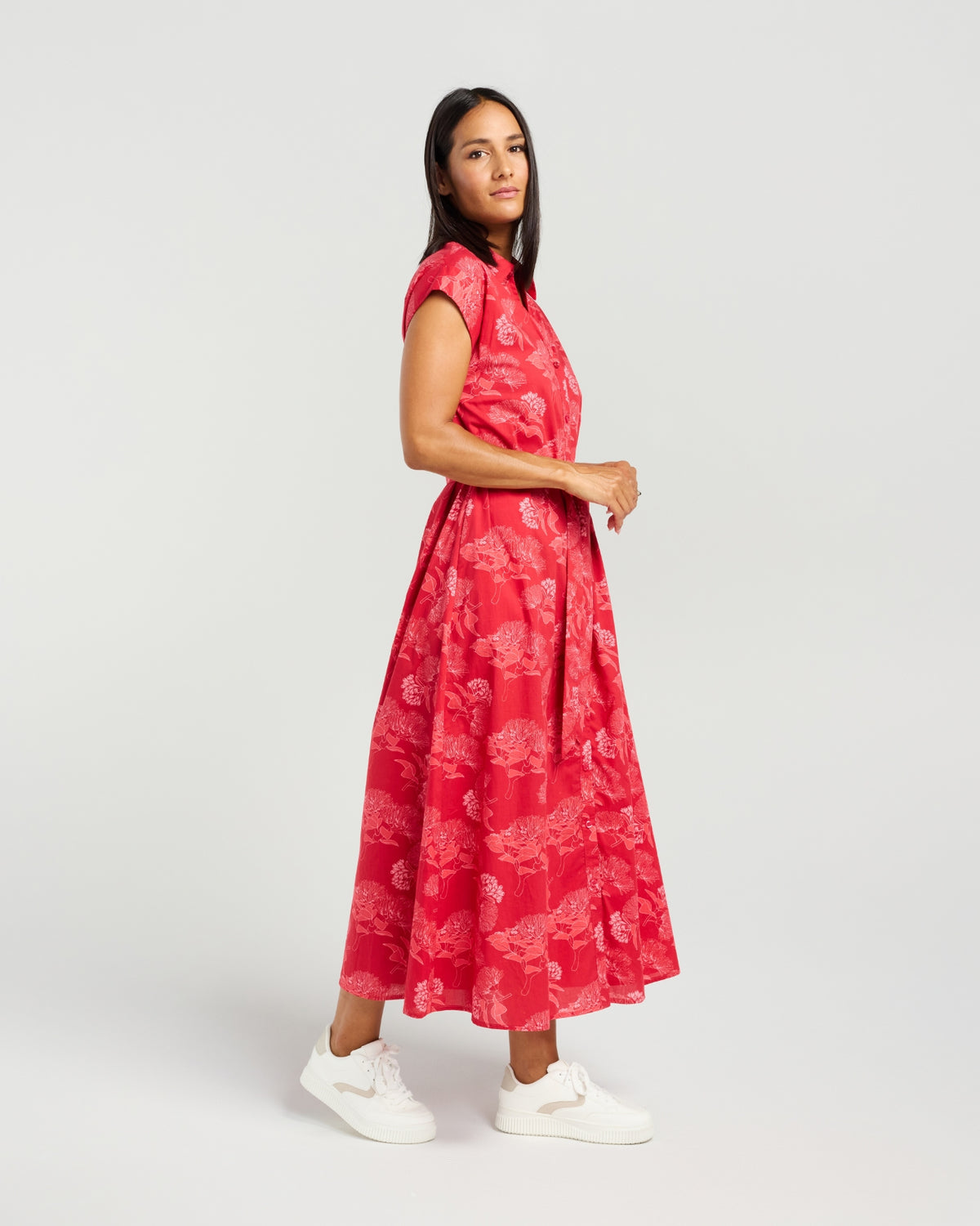 A woman modeling a red sleeveless midi dress with a tonal floral jacquard pattern, paired with white sneakers, against a light gray studio background.