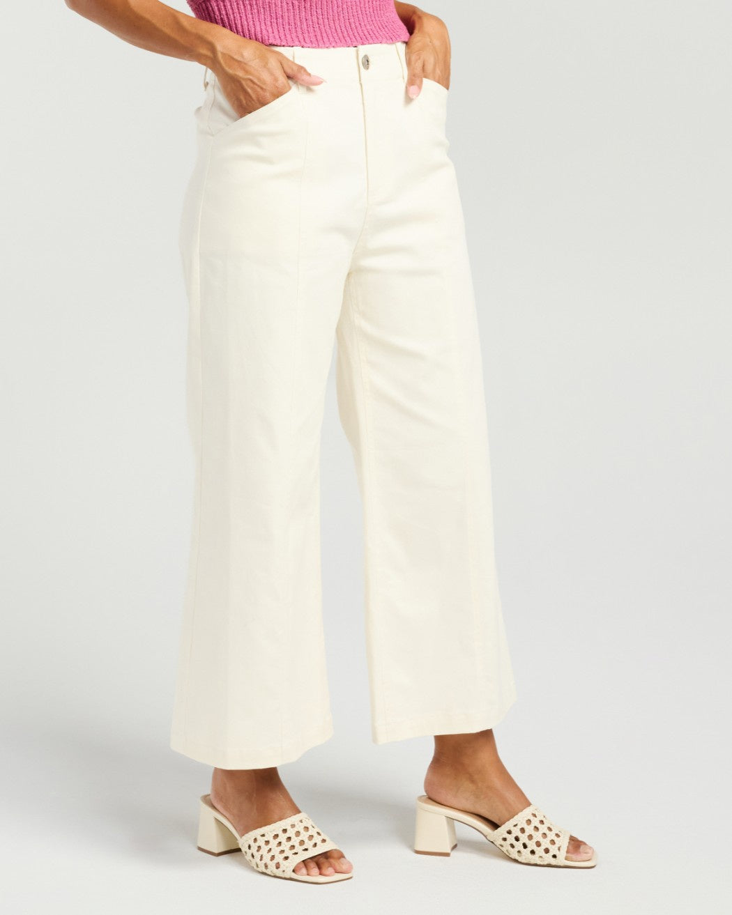 A model wearing off-white cropped wide-leg pants with a pink ribbed top and beige open-toe block-heel mules, shown from the waist down on a light gray studio background.