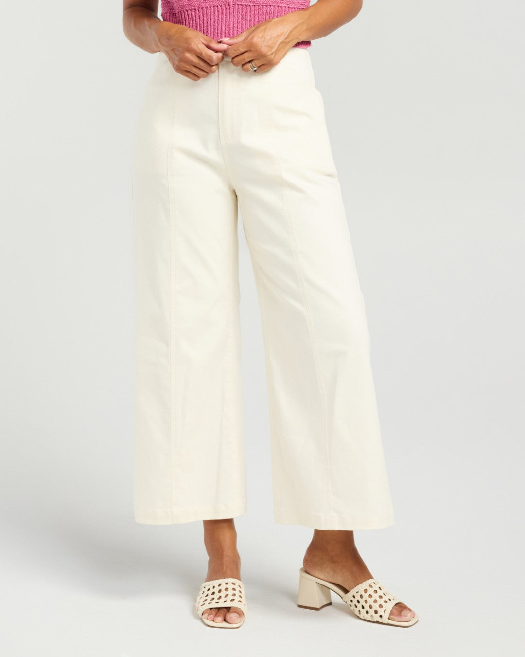 A model wearing off-white wide-leg cropped pants, a pink ribbed top, and cream-colored open-toe heeled sandals with cutout details, set against a plain light gray studio background.