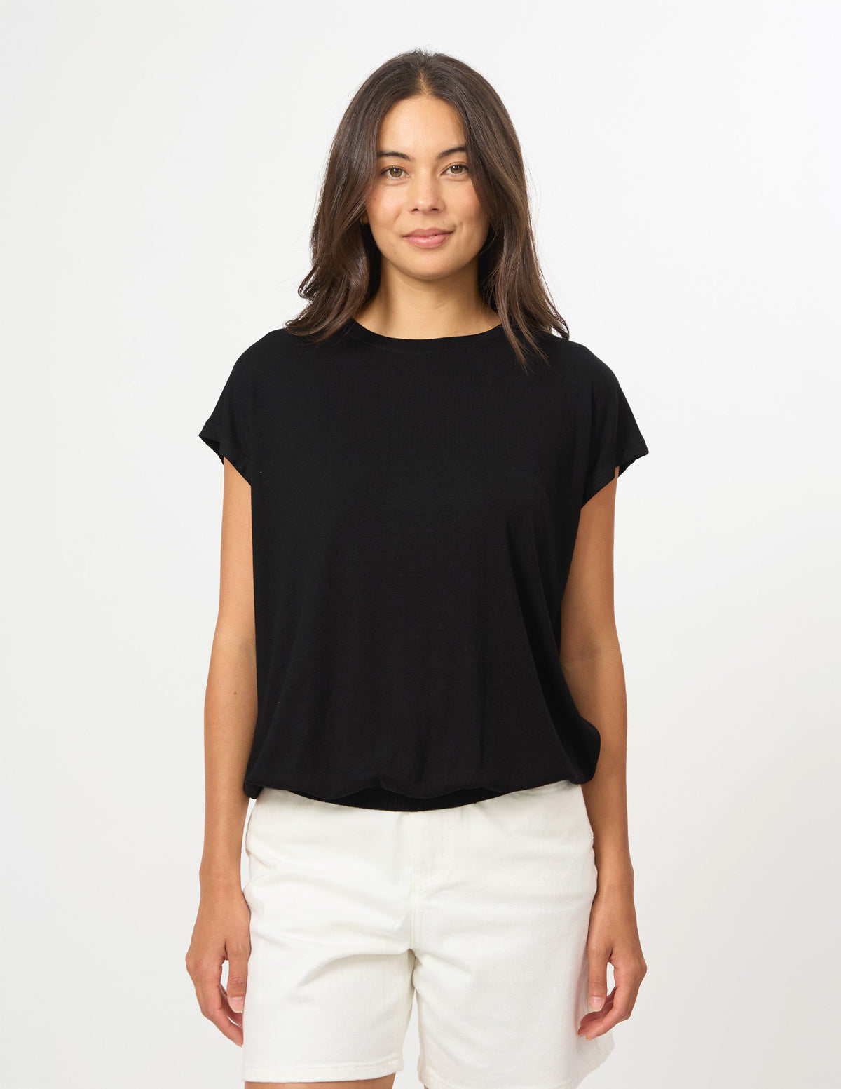 A woman modeling a black short-sleeve crewneck t-shirt paired with white shorts, standing against a plain white studio background.