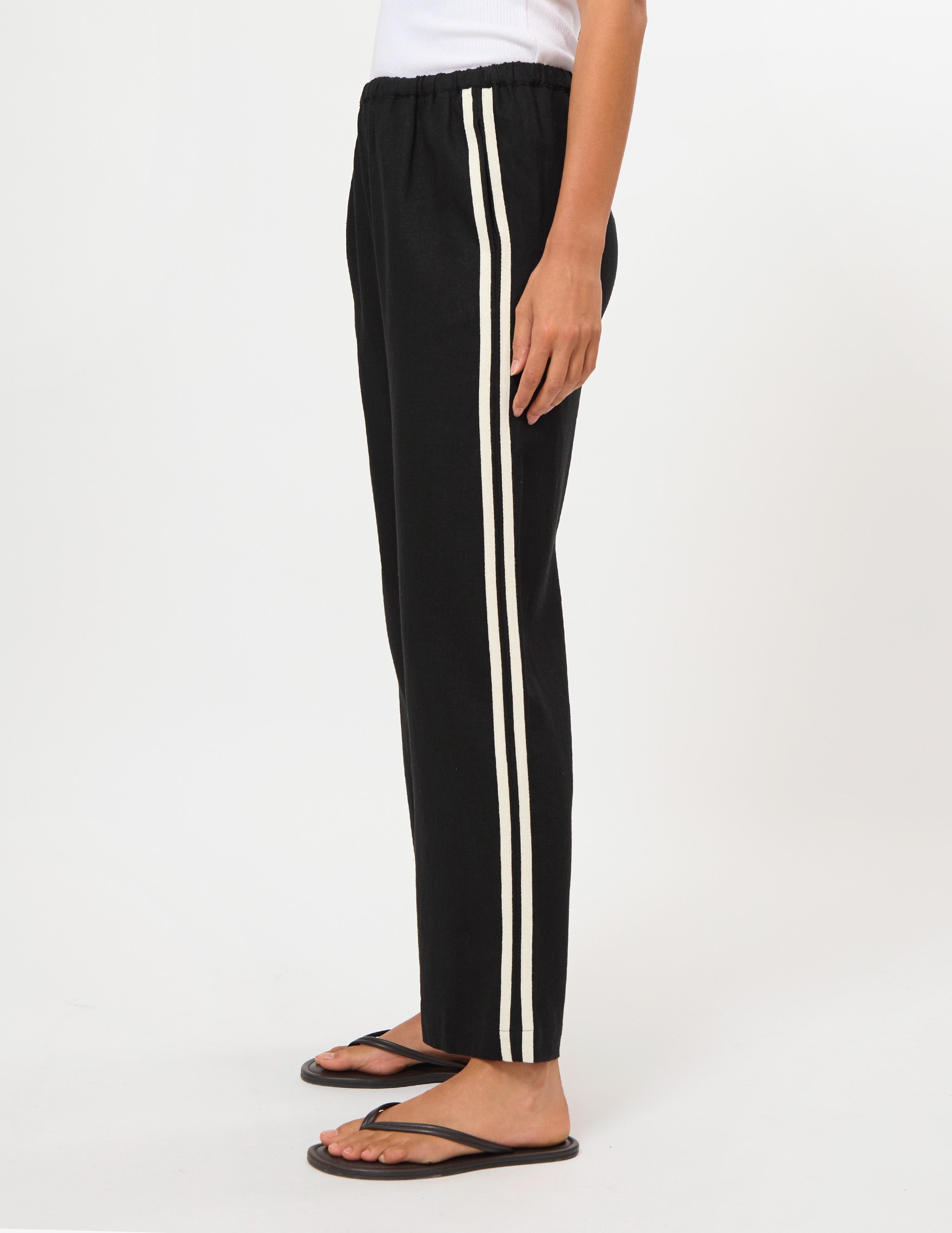 Black wide-leg track pants with two vertical cream stripes down the side, shown on a model wearing a white top and black flip-flops against a plain white studio background.