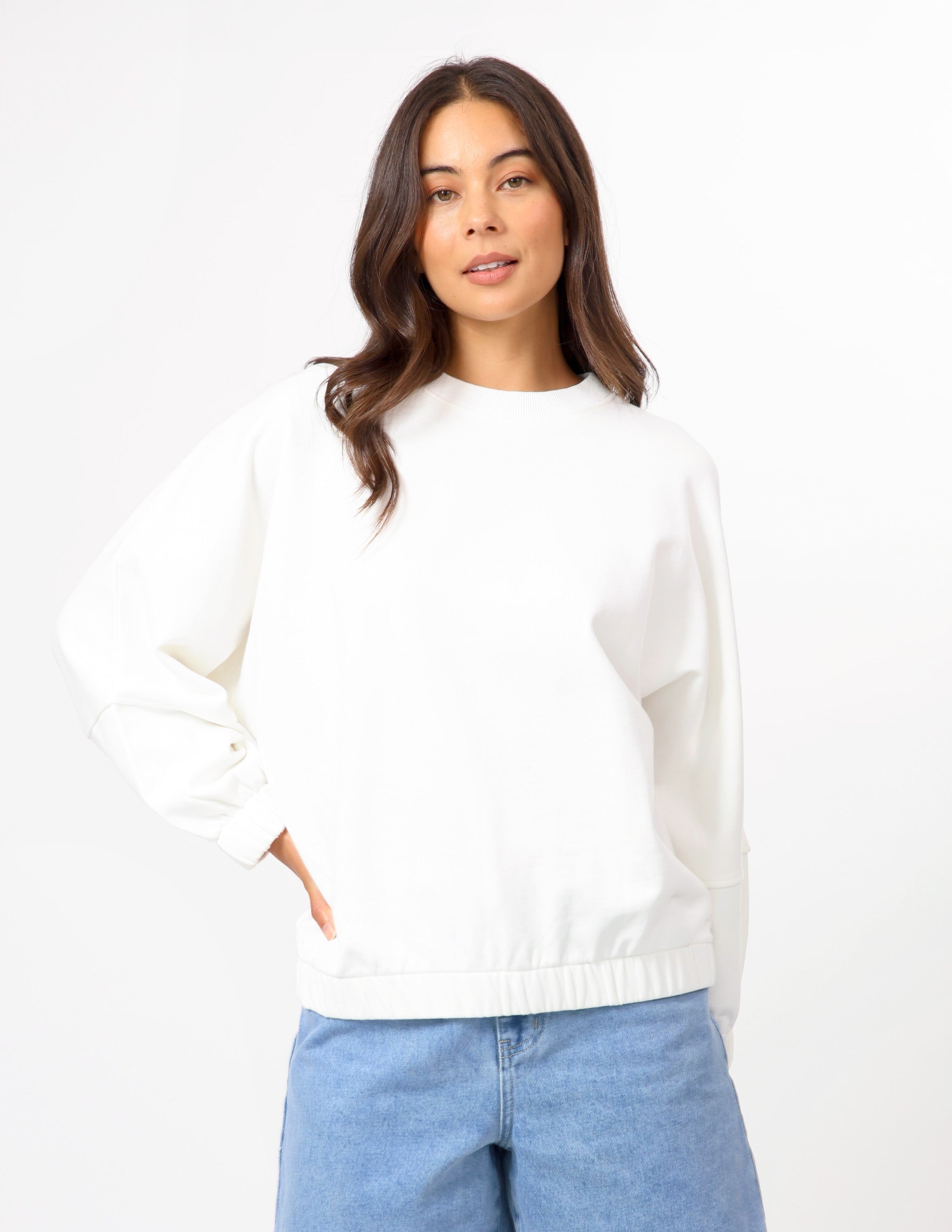 A woman wearing a white long-sleeve crewneck sweatshirt and light blue jeans, standing against a plain white studio background.