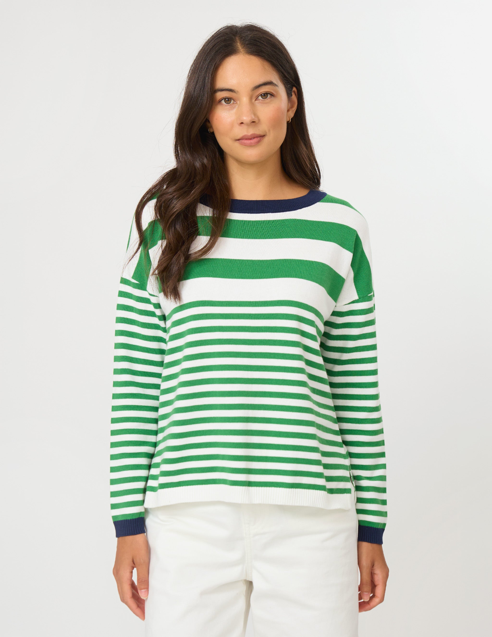 A woman wearing a green and white horizontally striped long-sleeve knit sweater with a navy blue crew neck, cuffs, and collar trim, paired with white pants, against a plain light gray studio background.