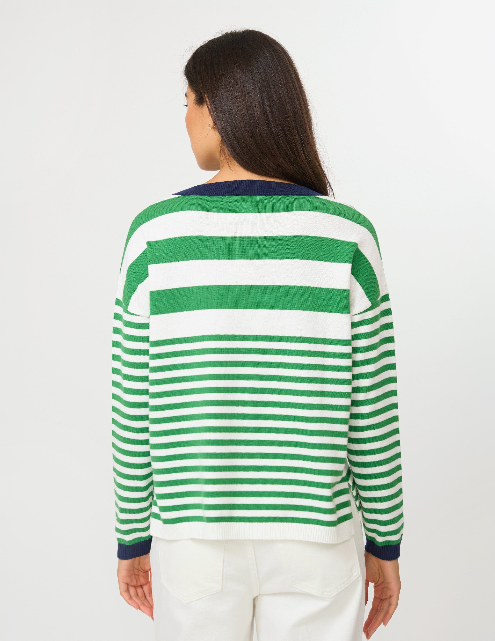 A woman viewed from the back wearing a green and white horizontally striped knit sweater with dark navy cuffs and collar, paired with white pants, against a plain light gray studio background.