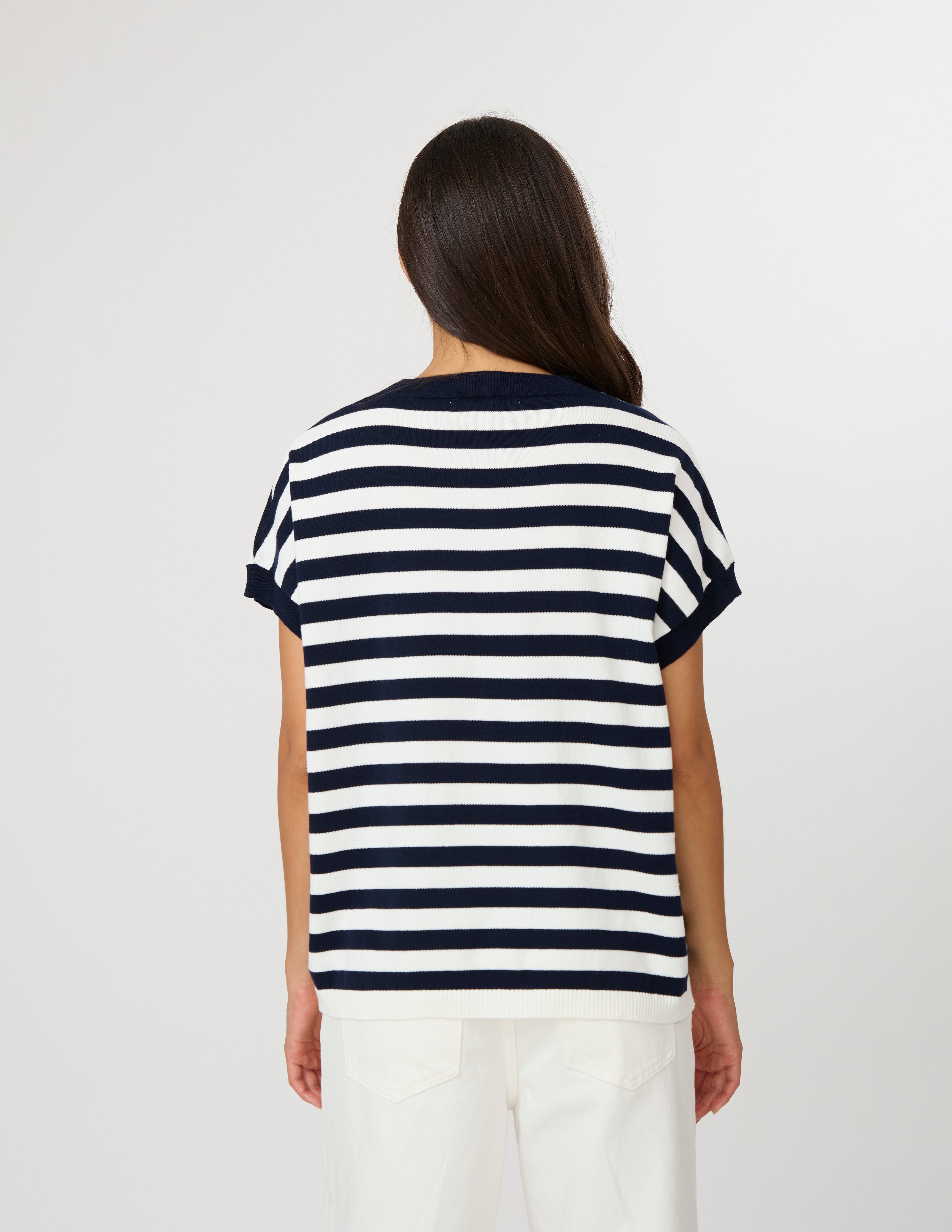 A woman viewed from the back wearing a short-sleeve striped T-shirt with navy and white horizontal stripes, paired with white pants, against a plain light gray studio background.
