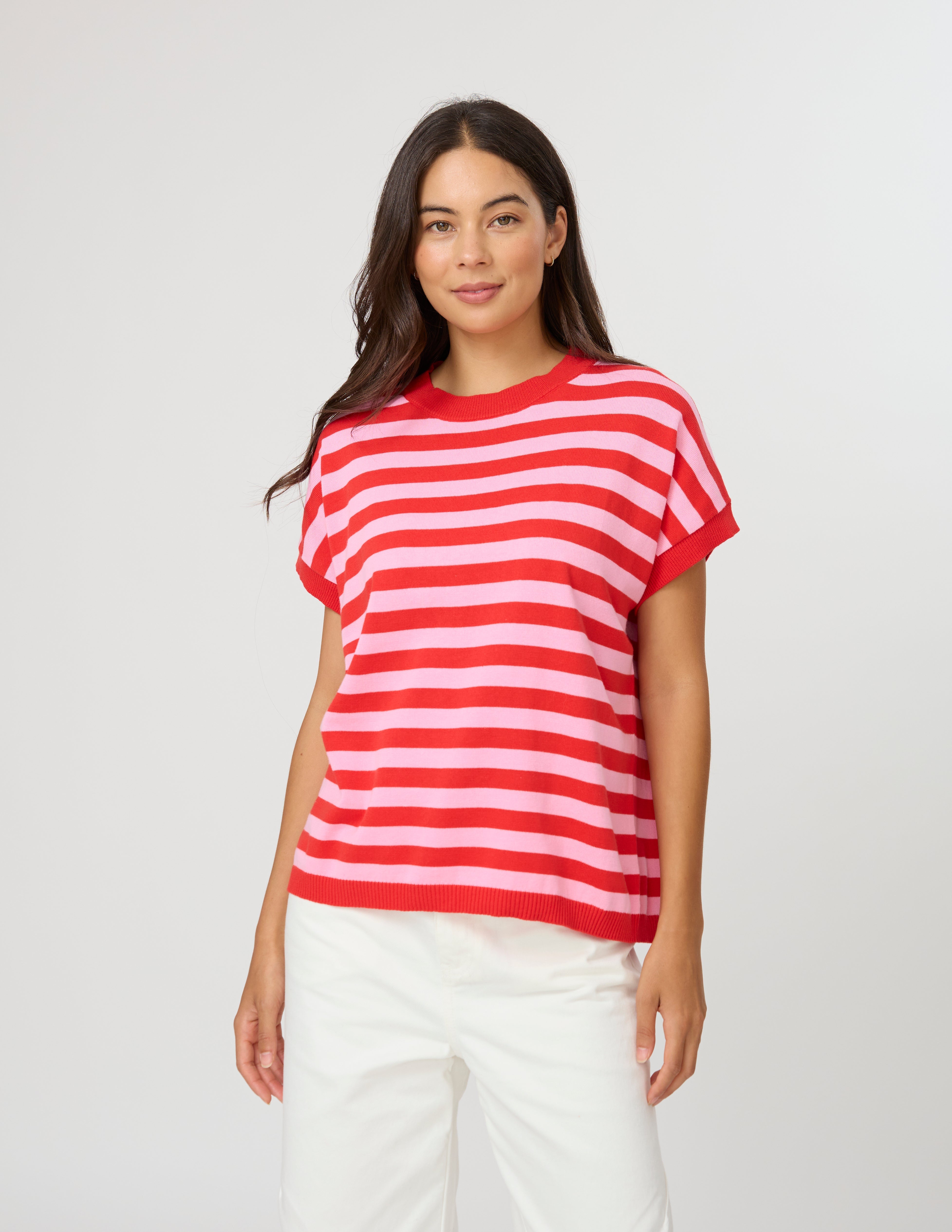 A woman modeling a red and white horizontally striped short-sleeve knit top with white pants, standing against a light gray studio background.