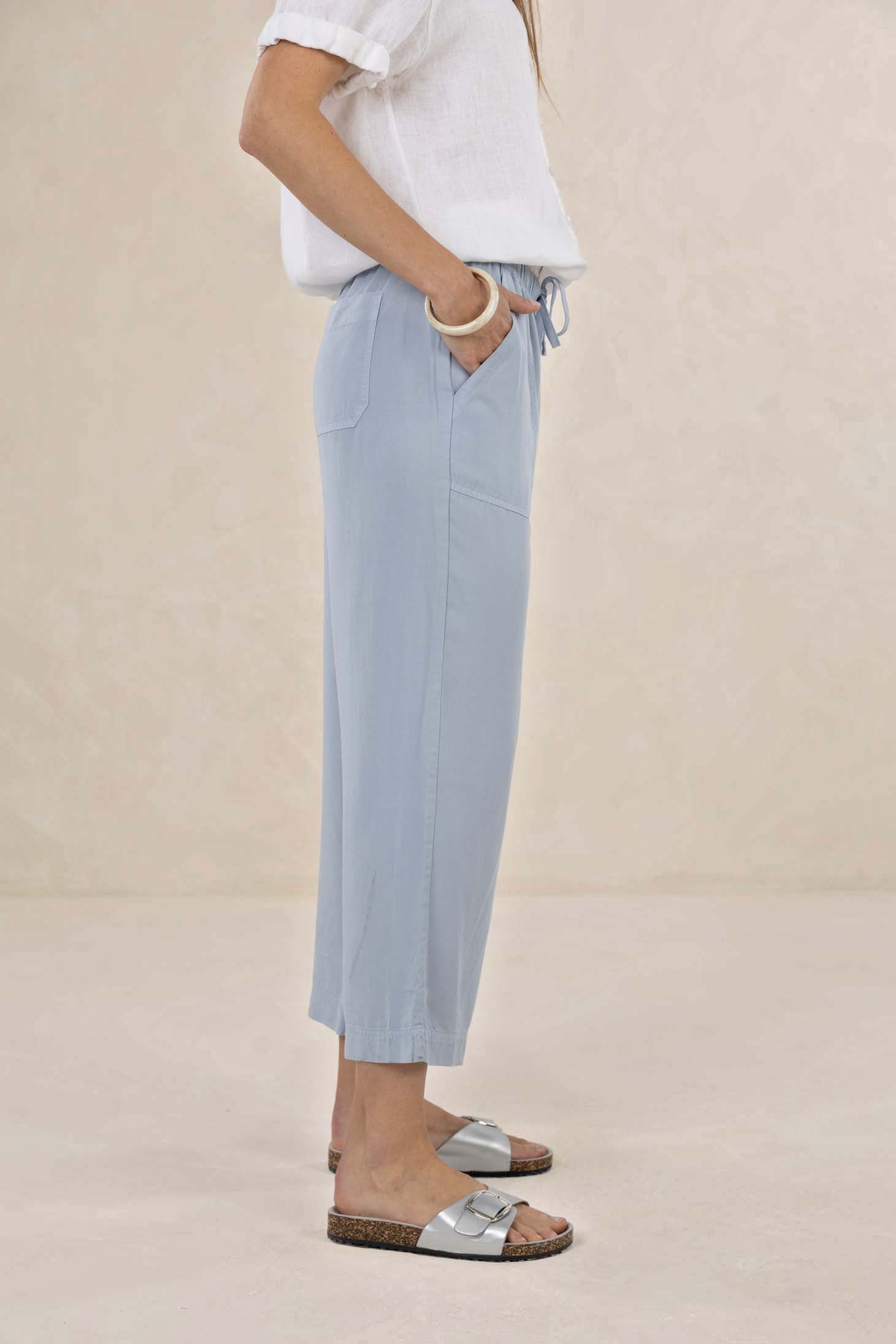 A model wearing light blue wide-leg cropped pants with a drawstring waist, paired with a white top and silver slide sandals, standing against a neutral beige background.