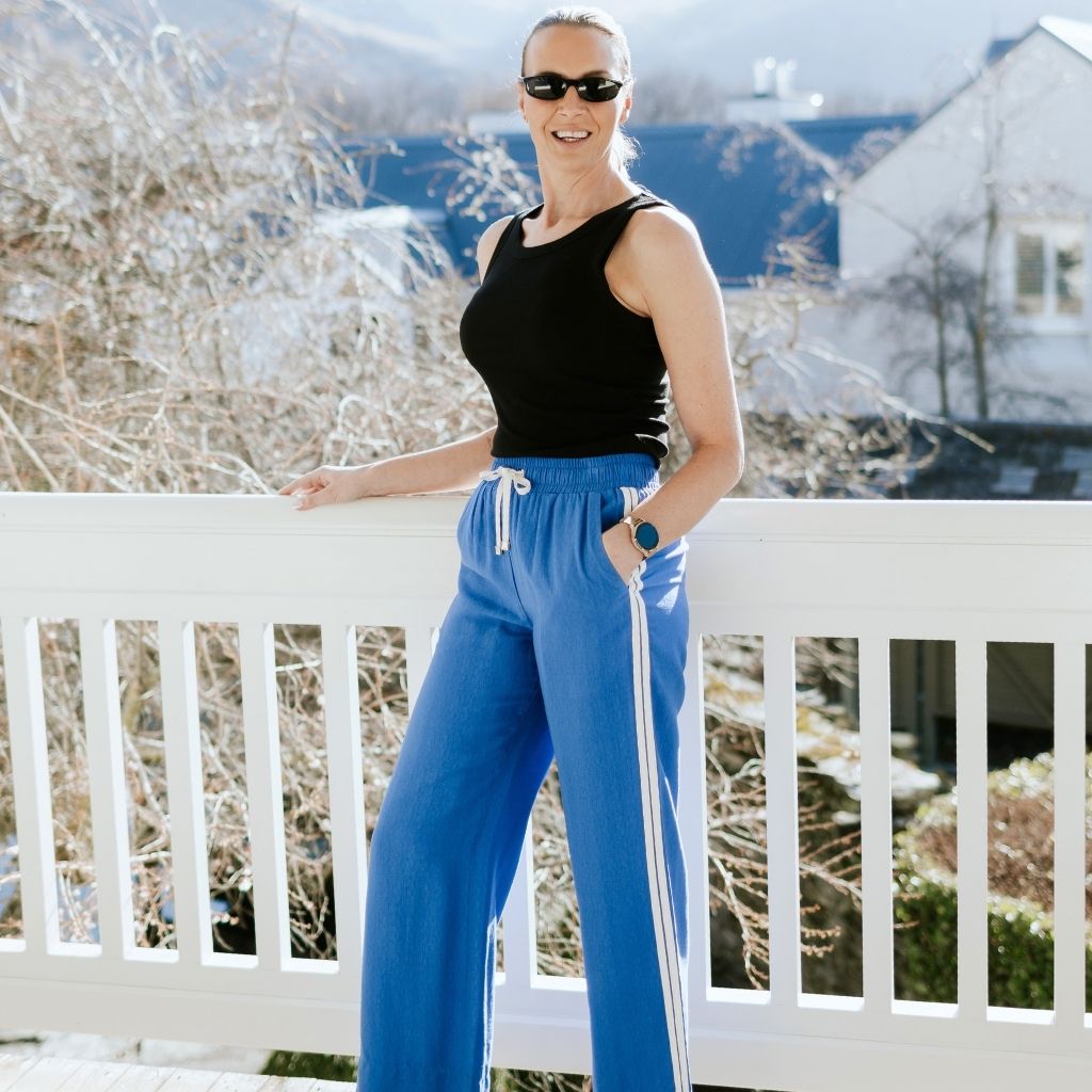 A woman wearing blue wide-leg athletic pants with white side stripes, a black sleeveless top, and sunglasses, posing outdoors on a white balcony railing with houses and trees in the background.