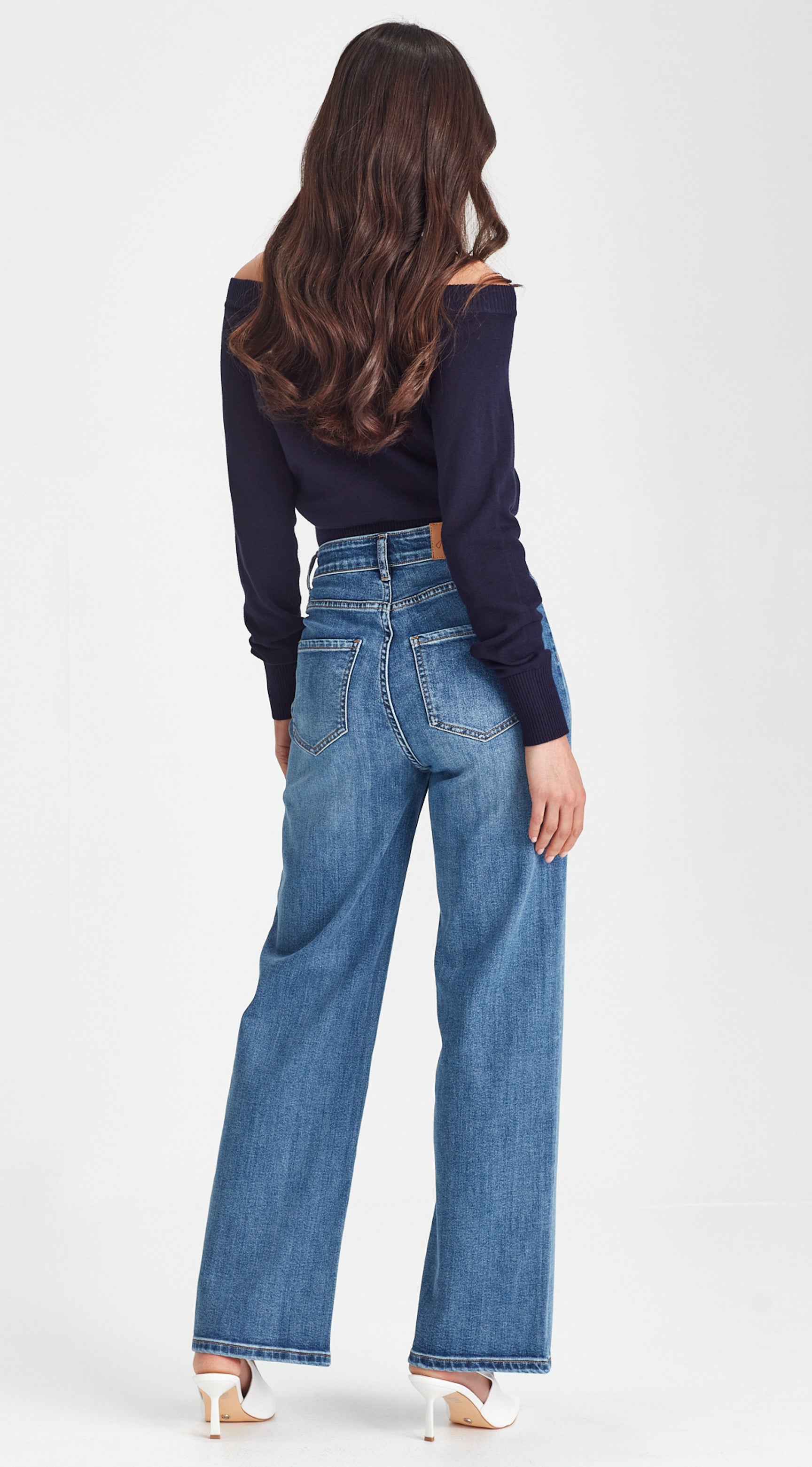 A woman shown from the back wearing high-waisted, wide-leg blue denim jeans and a dark navy off-the-shoulder long-sleeve top, paired with white high-heeled sandals, against a plain white studio background.