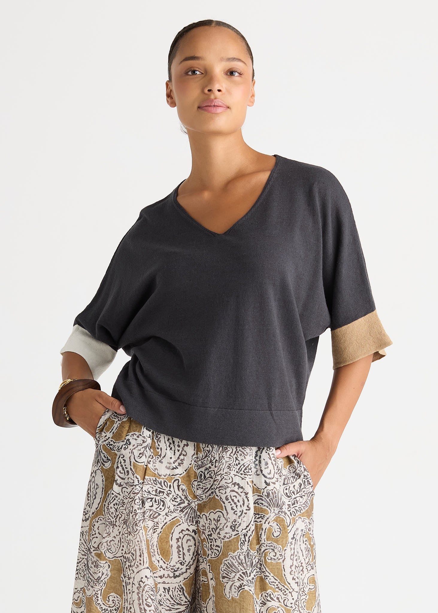 A woman modeling a dark charcoal gray V-neck knit top with contrasting beige sleeve cuffs, paired with patterned trousers, against a plain light gray studio background.