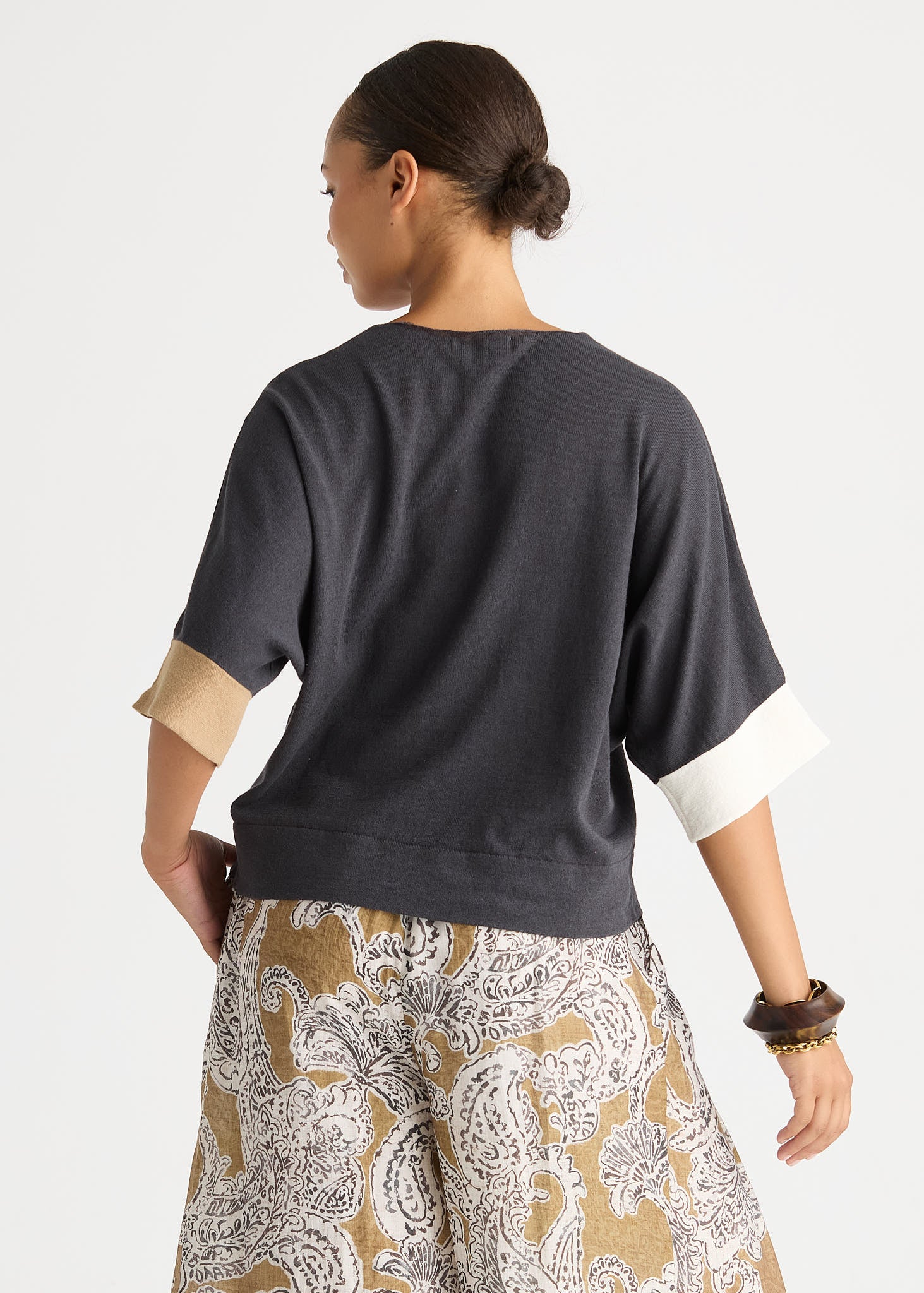 A woman shown from the back wearing a dark charcoal gray short-sleeve top with contrasting beige and white rolled cuffs, paired with patterned beige and white pants, against a light gray studio background.