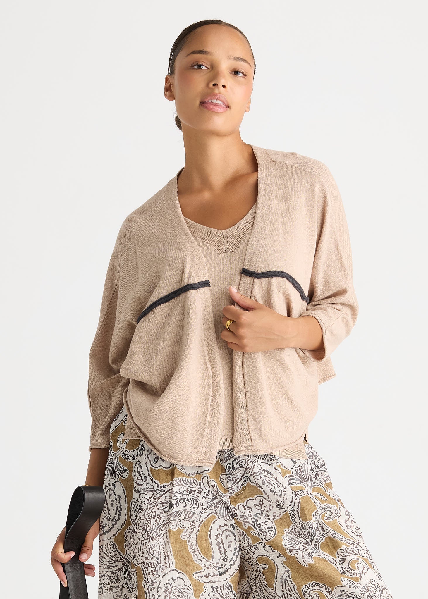 A woman modeling a light beige open-front cardigan with black piping details over a matching beige knit top, paired with a patterned skirt, against a plain light gray background.